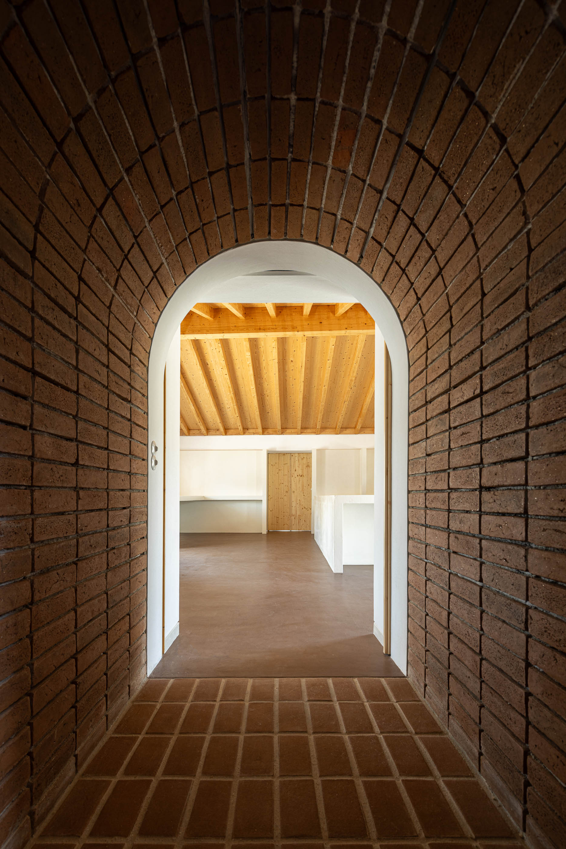 Corredor: Arco de tijolo que emoldura a entrada para um espaço interior com pavimento castanho e teto de madeira. — Olaria Municipal arquitetura ARDE Arquitetura+Design em Odemira