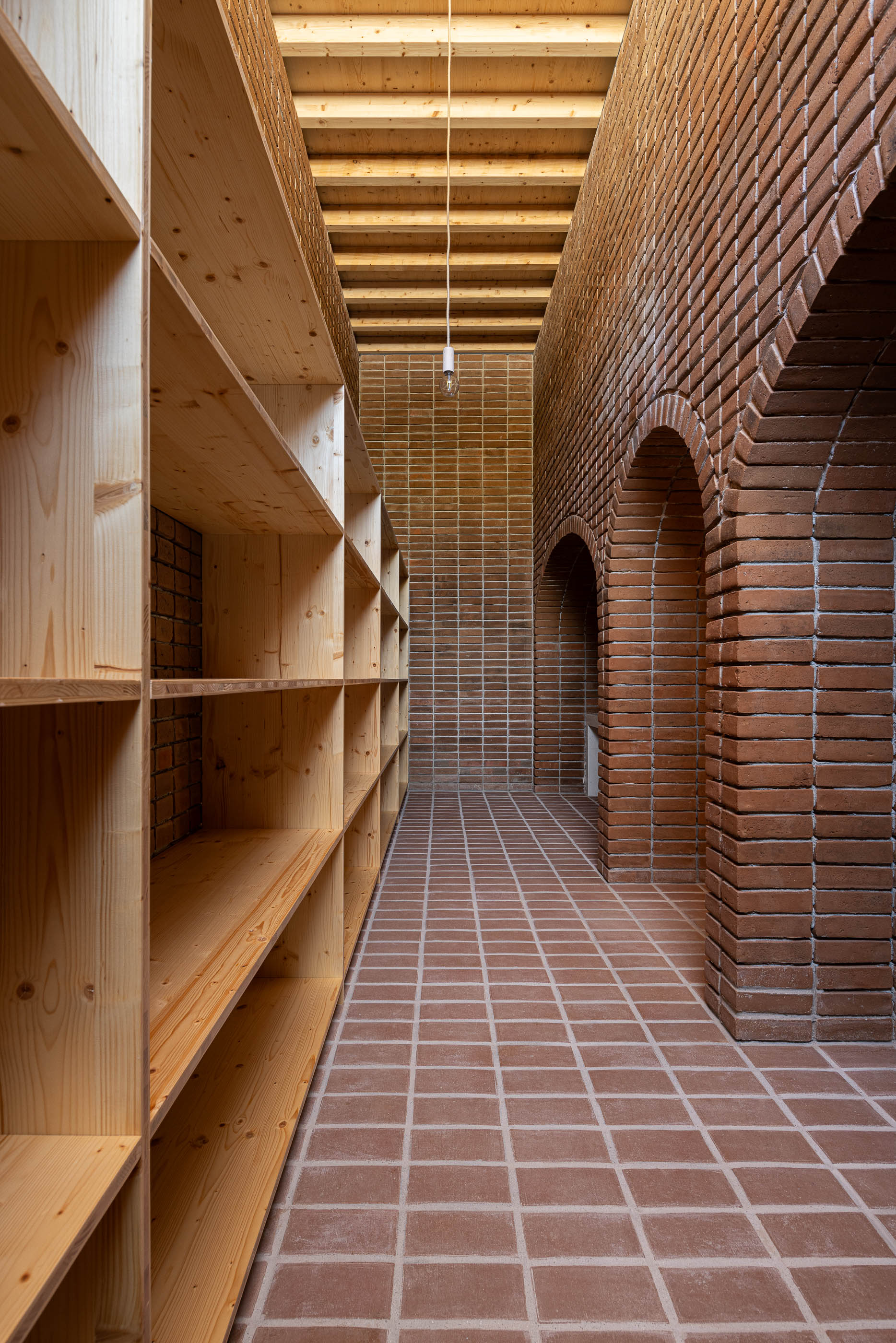 Corredor: Corredor estreito com prateleiras de madeira, paredes de tijolo e teto de madeira com claraboia. — Olaria Municipal arquitetura ARDE Arquitetura+Design em Odemira