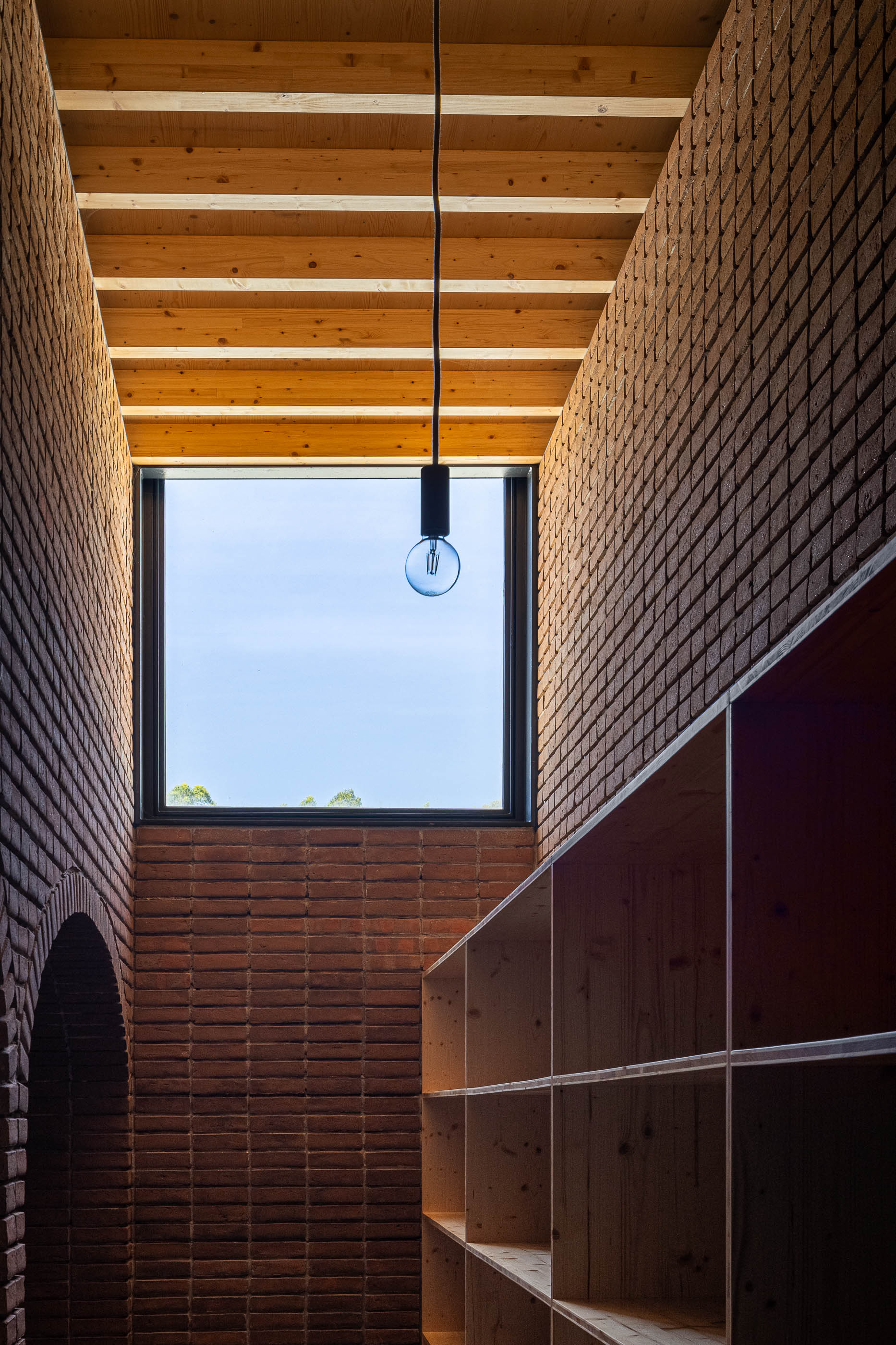 Corredor: Claraboia com teto de madeira e paredes de tijolo, com prateleiras de madeira. — Olaria Municipal arquitetura ARDE Arquitetura+Design em Odemira