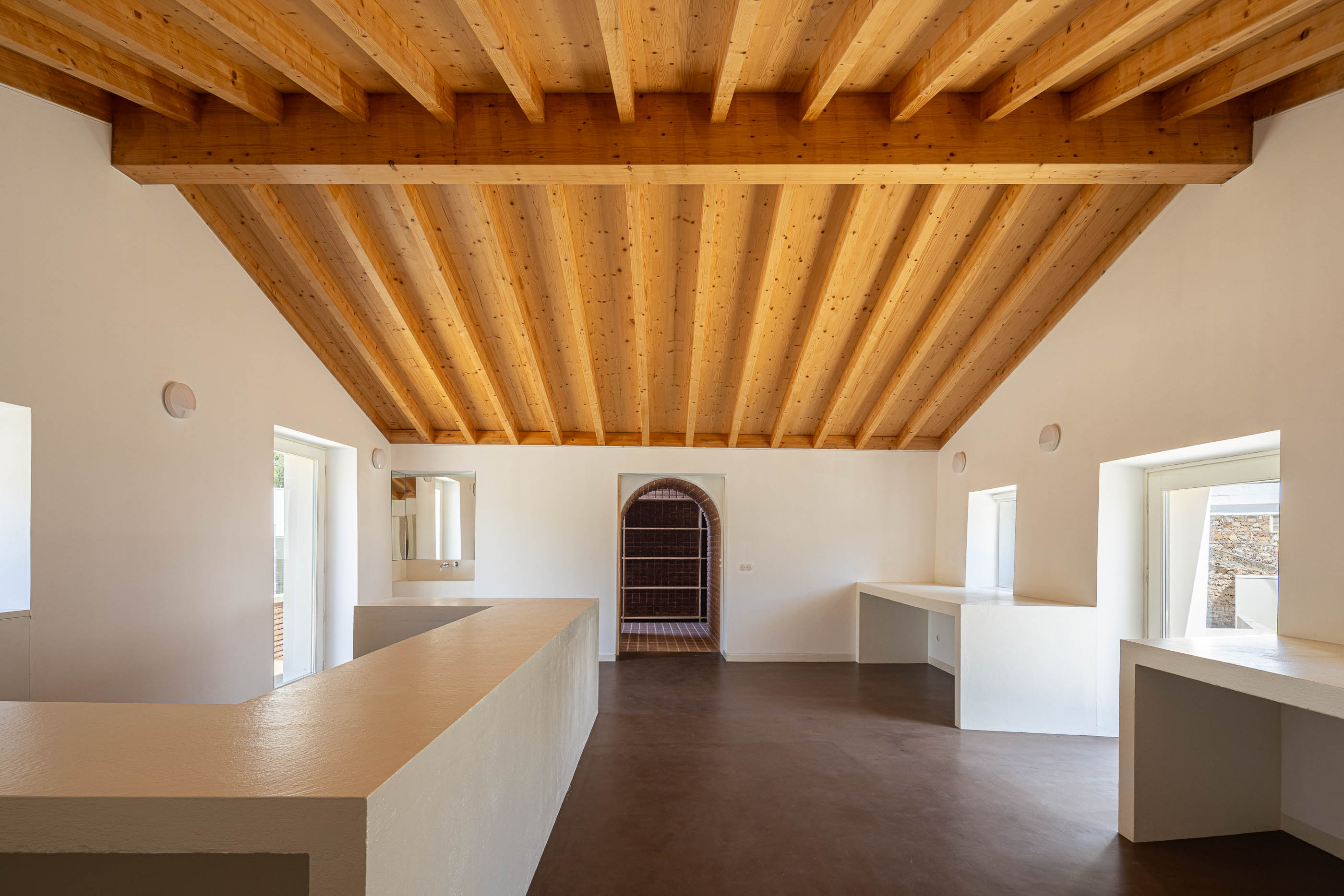 Espaço de trabalho: Sala ampla com teto de madeira, pavimento castanho, bancadas brancas e acesso ao corredor de tijolo. — Olaria Municipal arquitetura ARDE Arquitetura+Design em Odemira
