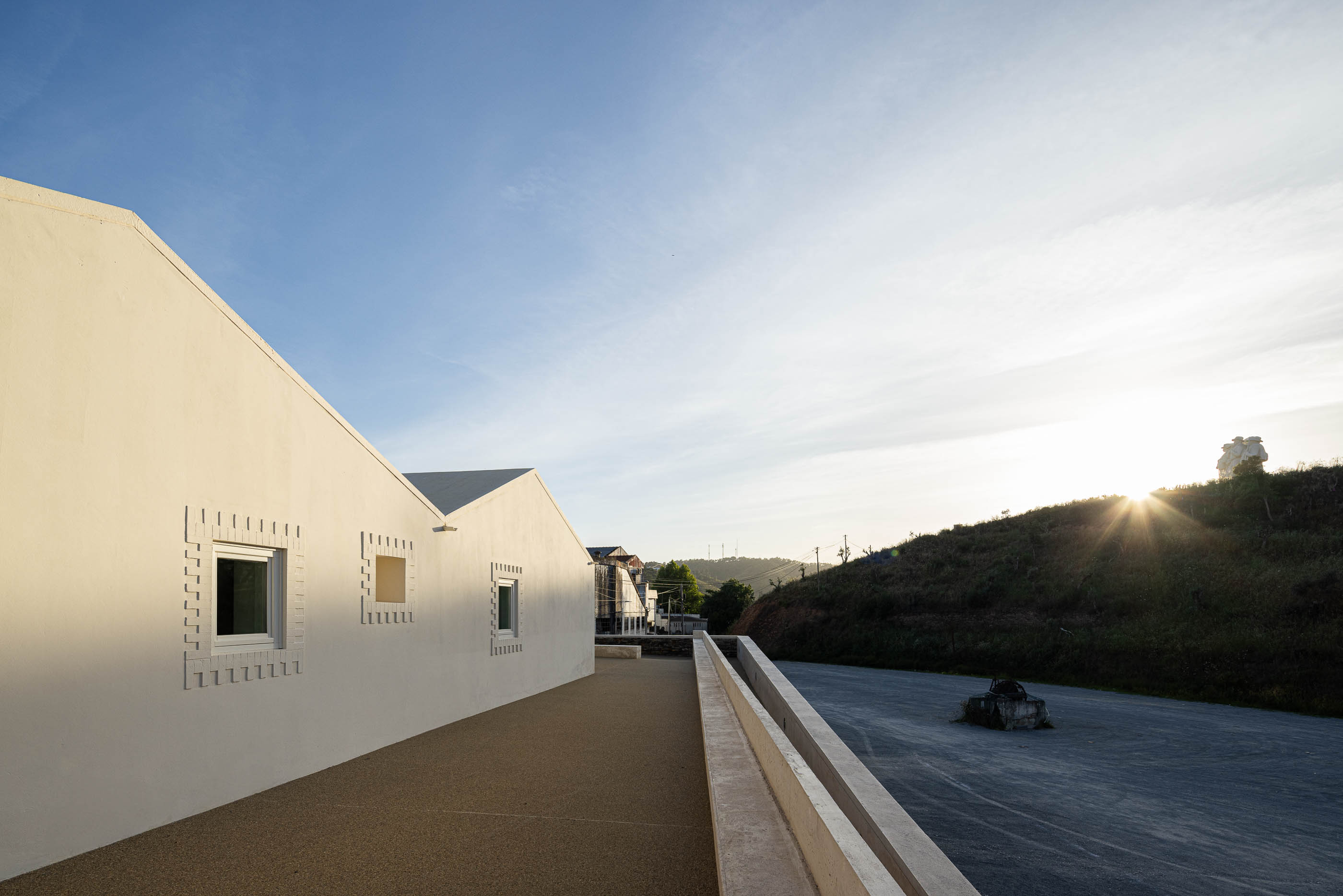 Exterior: Vista lateral do edifício branco com detalhes em tijolo e paisagem ao fundo. — Olaria Municipal arquitetura ARDE Arquitetura+Design em Odemira