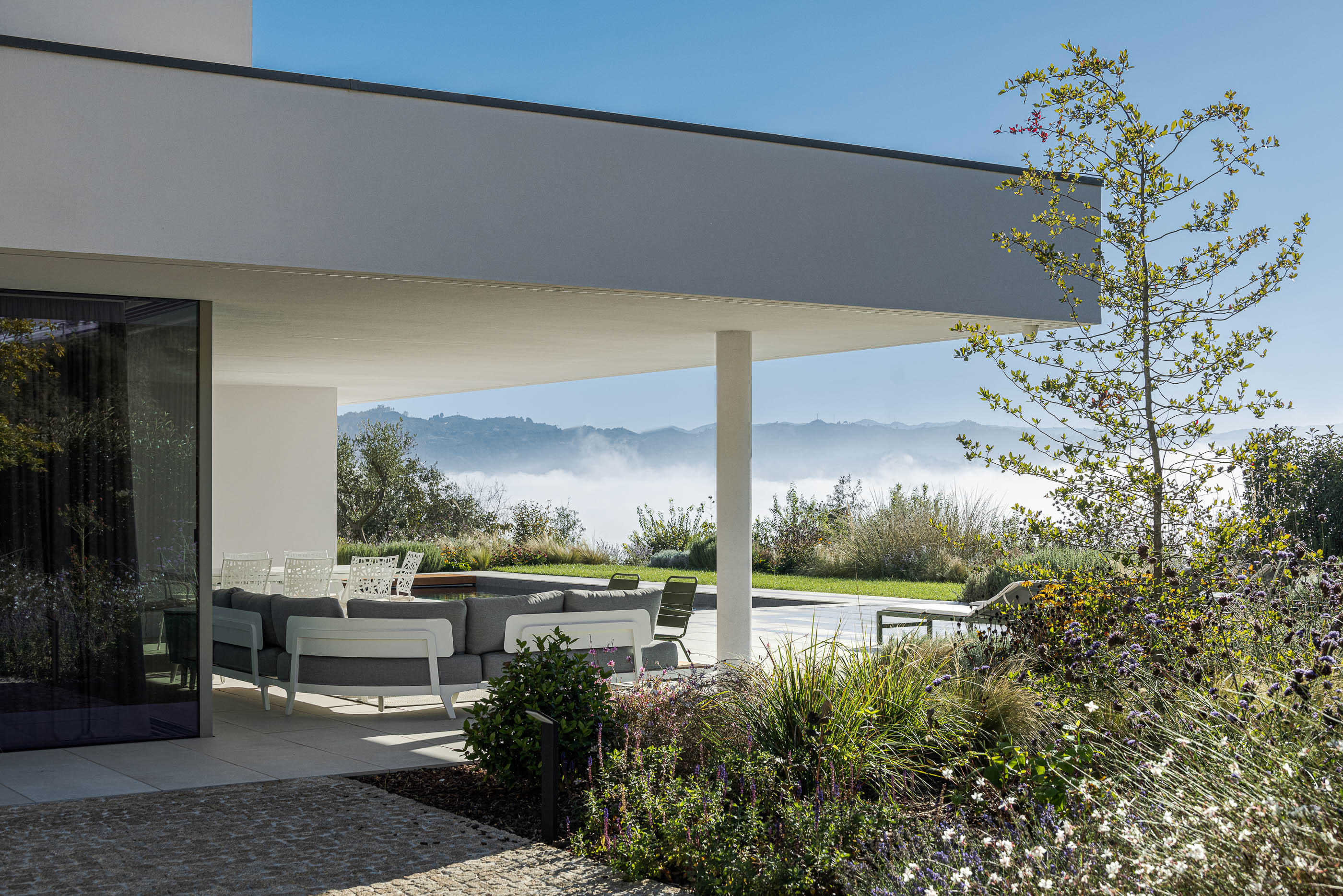 Terraço coberto com mobiliário de exterior cinzento e vista para a paisagem montanhosa envolta em neblina. — Habitação Áurea arquitetura stu.dere em Amarante