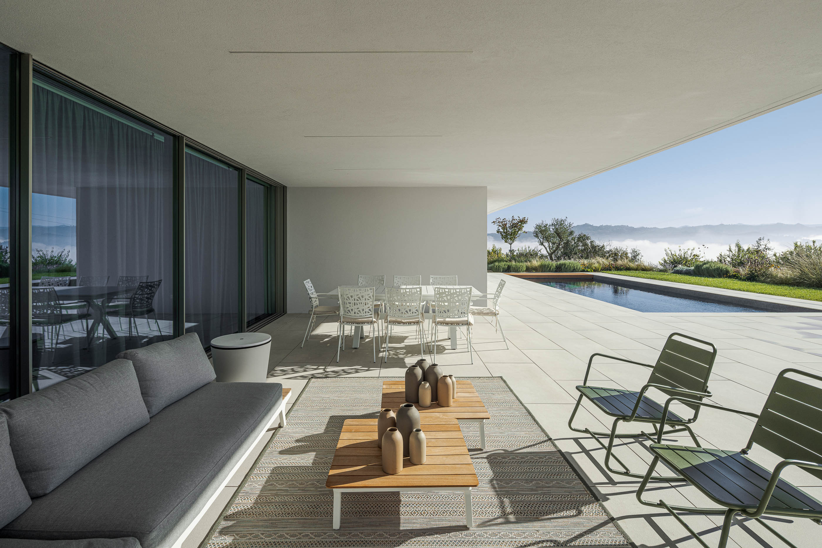 Terraço coberto com mesa de jantar branca, sofás cinzentos e alcatifa geométrica. — Habitação Áurea arquitetura stu.dere em Amarante