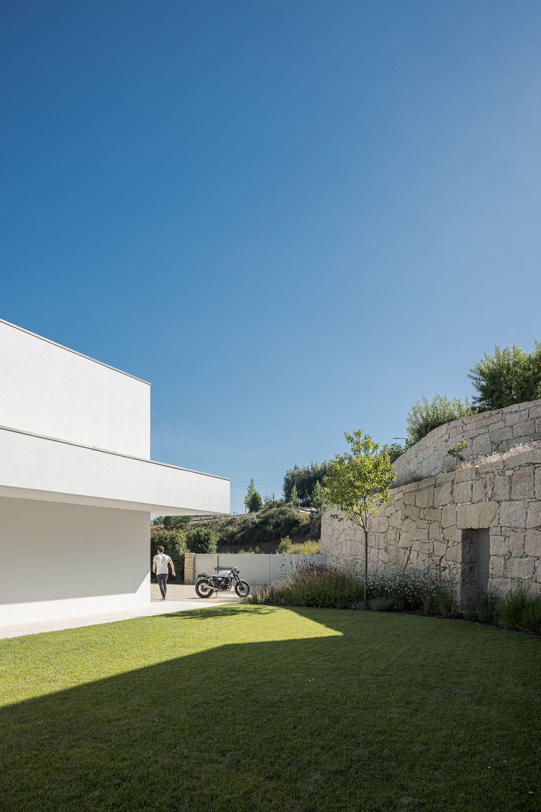 Exterior da moradia com relvado, muro de pedra e mota estacionada. — Habitação Áurea arquitetura stu.dere em Amarante