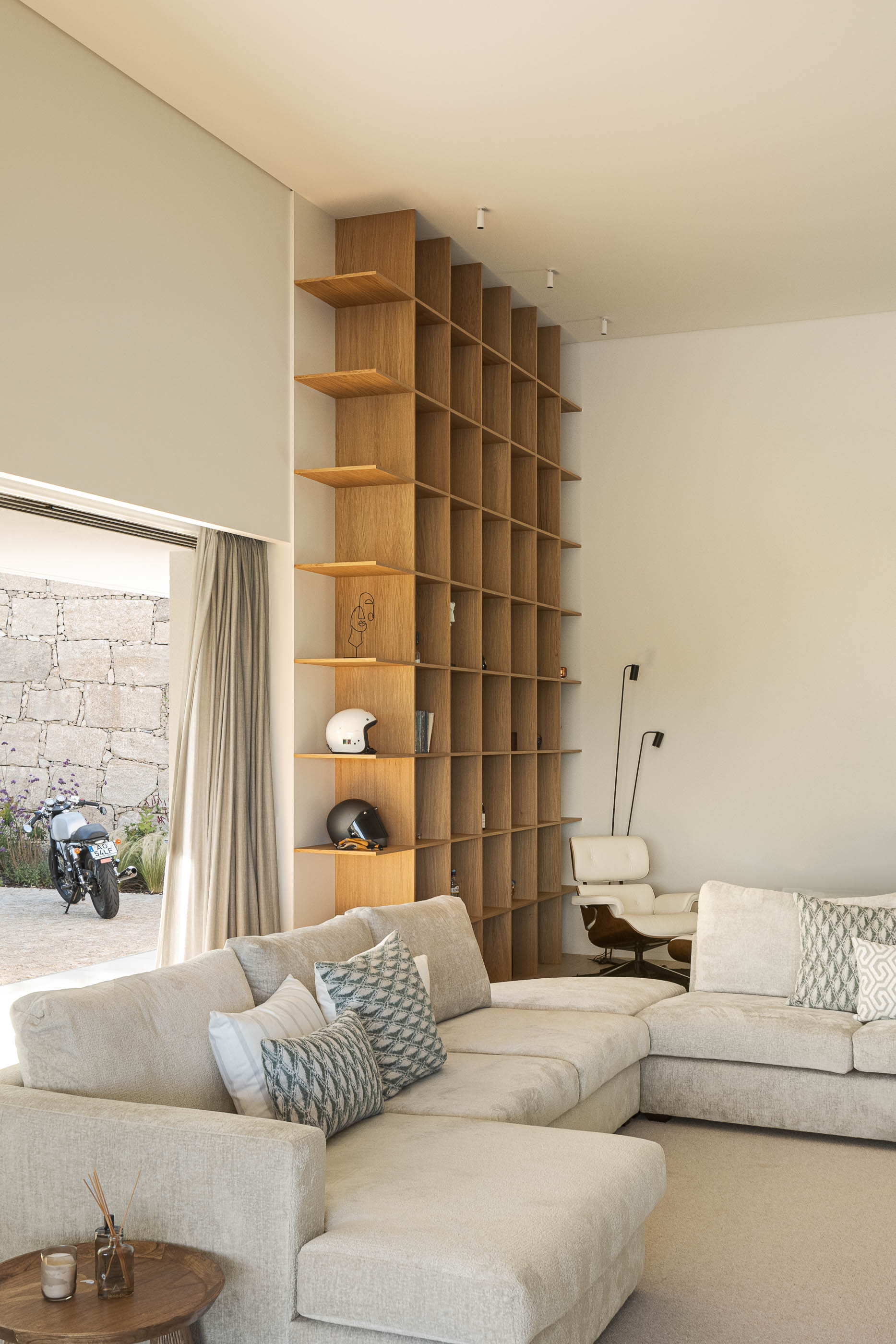 Sala de estar com estante de madeira, sofá bege e vista para o exterior. — Habitação Áurea arquitetura stu.dere em Amarante