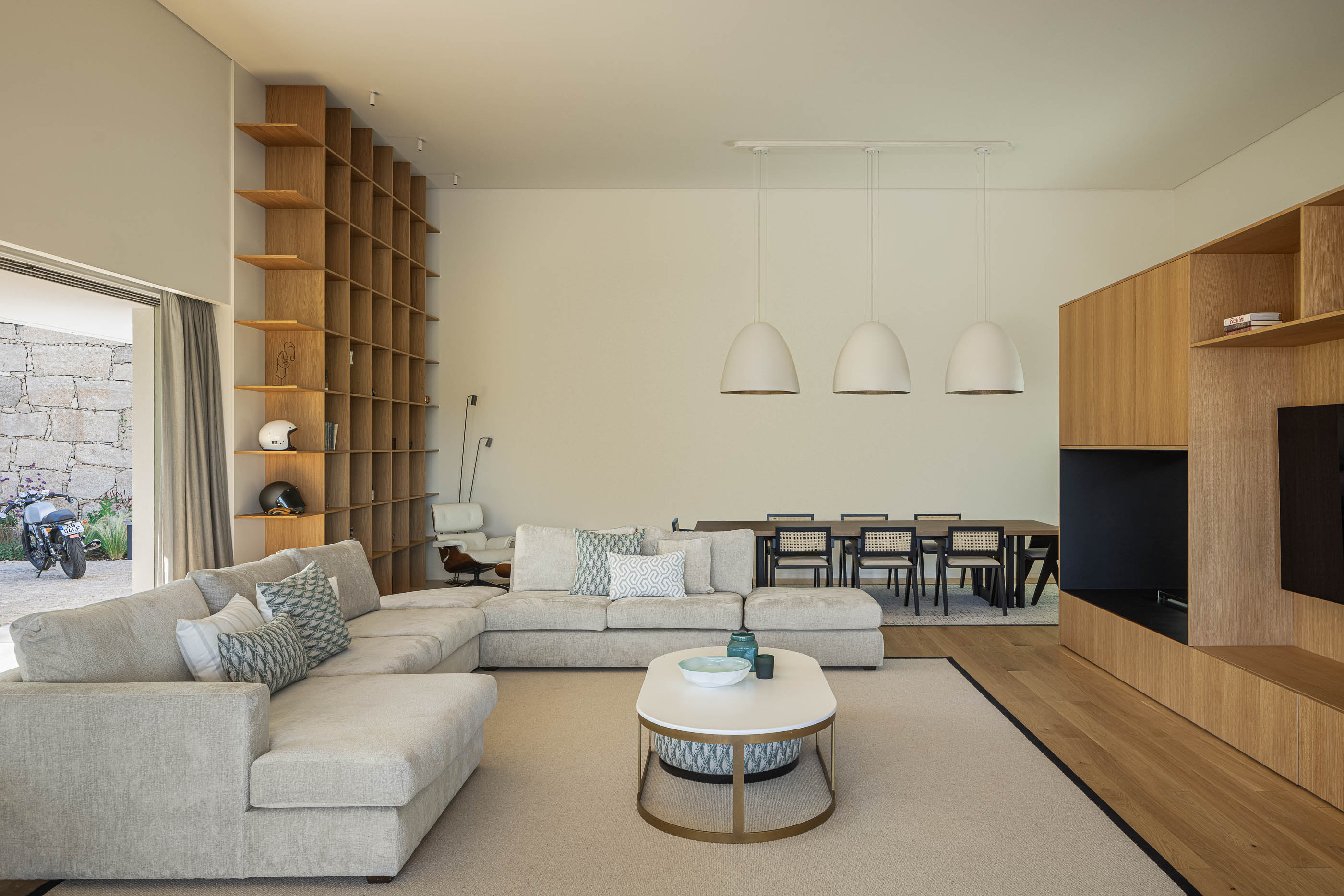 Sala de estar com sofá bege, estante de madeira e vista para a sala de jantar. — Habitação Áurea arquitetura stu.dere em Amarante
