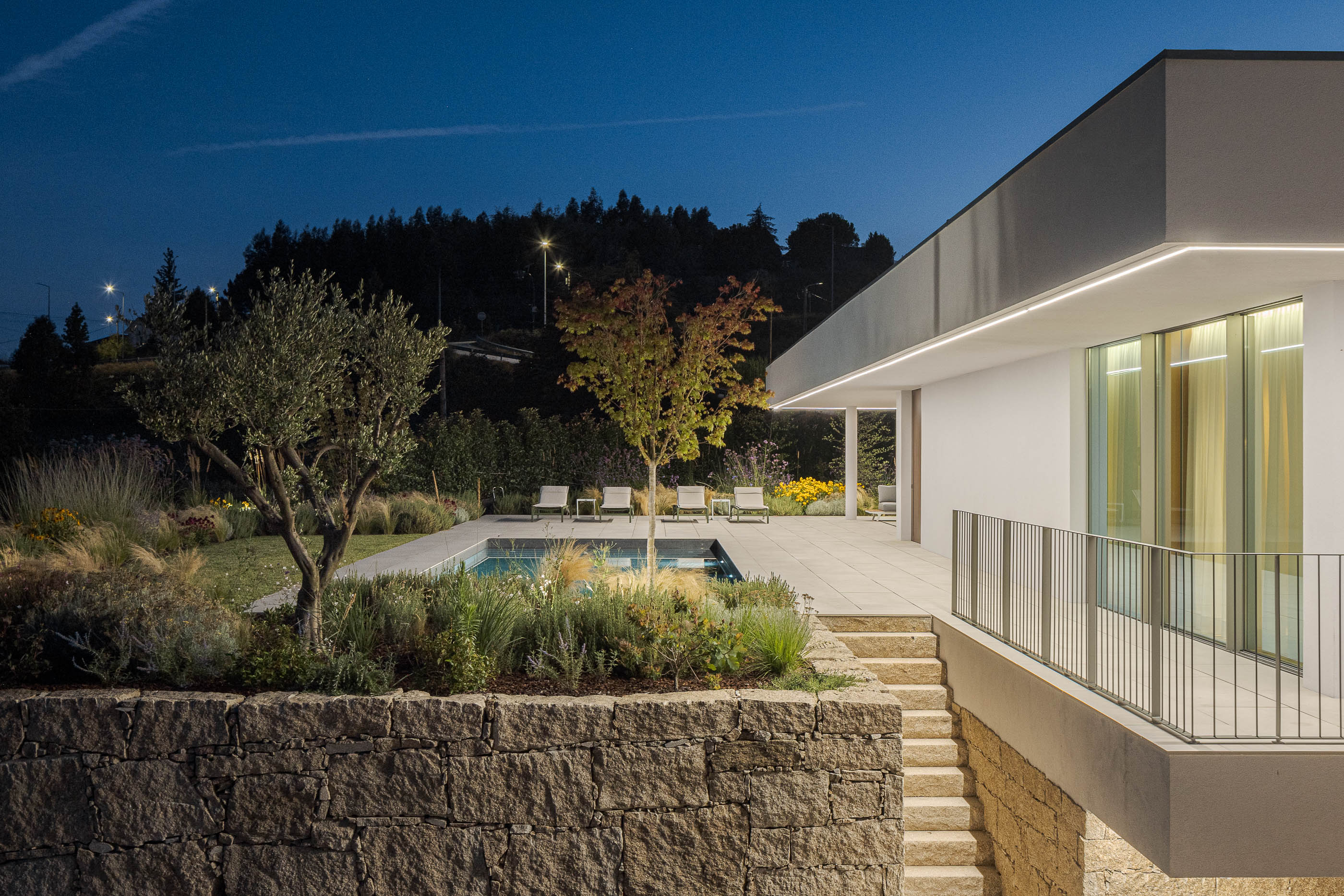 Exterior da moradia iluminada com piscina, jardim e muro de pedra. — Habitação Áurea arquitetura stu.dere em Amarante
