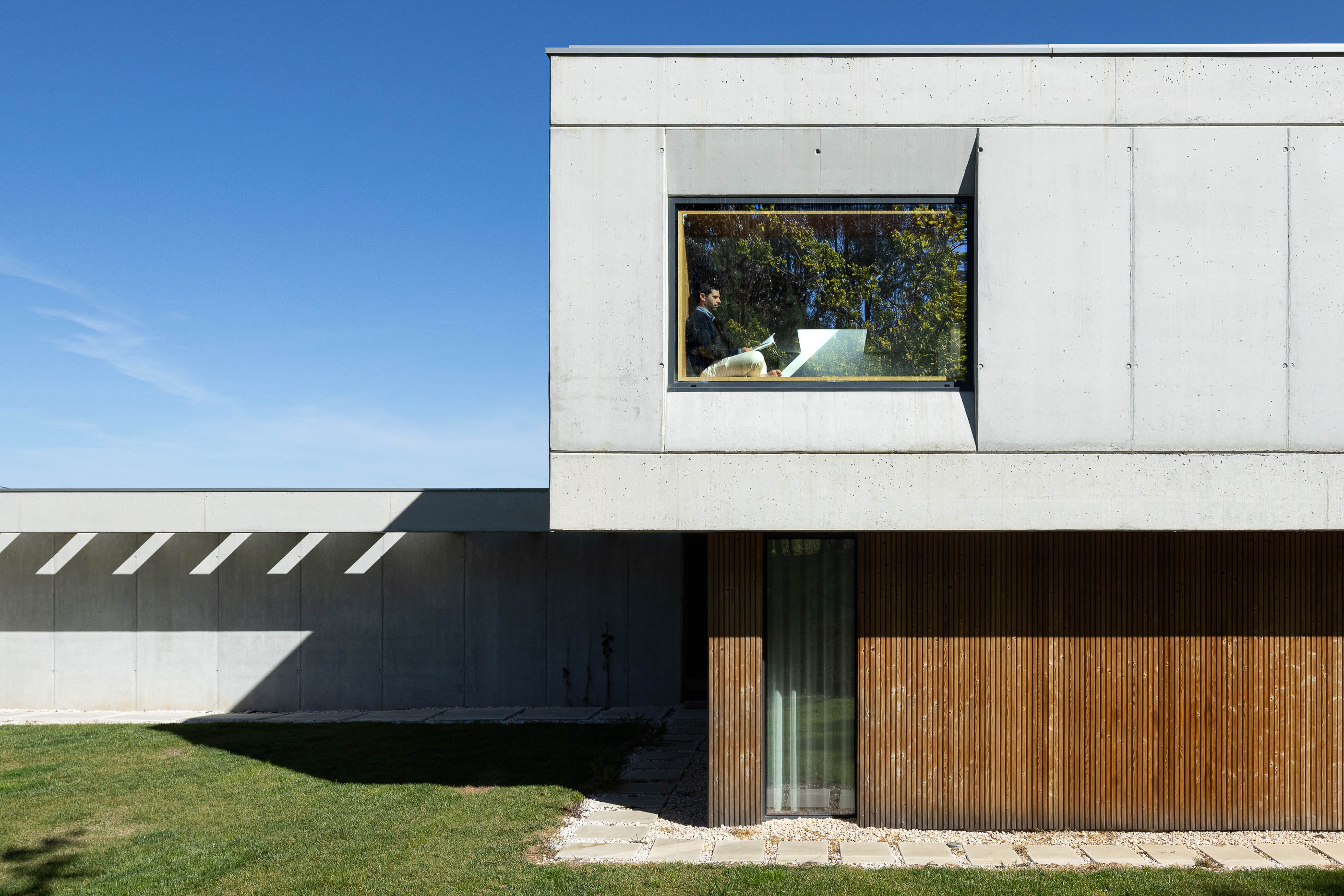 Quarto: Detalhe da fachada em betão e madeira com janela quadrada e pessoa a ler no interior. — Casa em Mundão arquitetura Um para Um, arquitectura e engenharia em Viseu