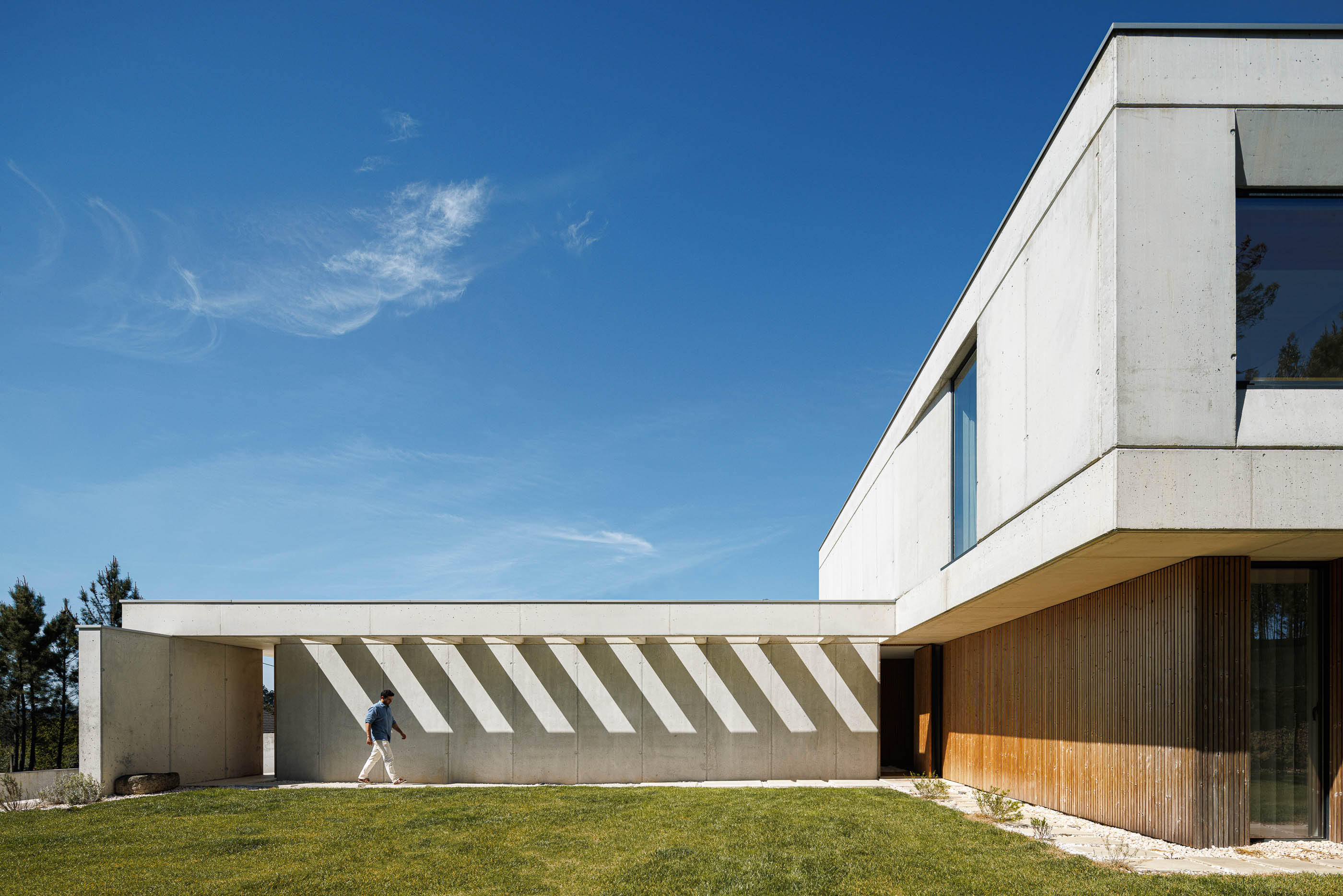 Entrada: Fachada da casa em betão e madeira com alpendre e homem a caminhar. — Casa em Mundão arquitetura Um para Um, arquitectura e engenharia em Viseu