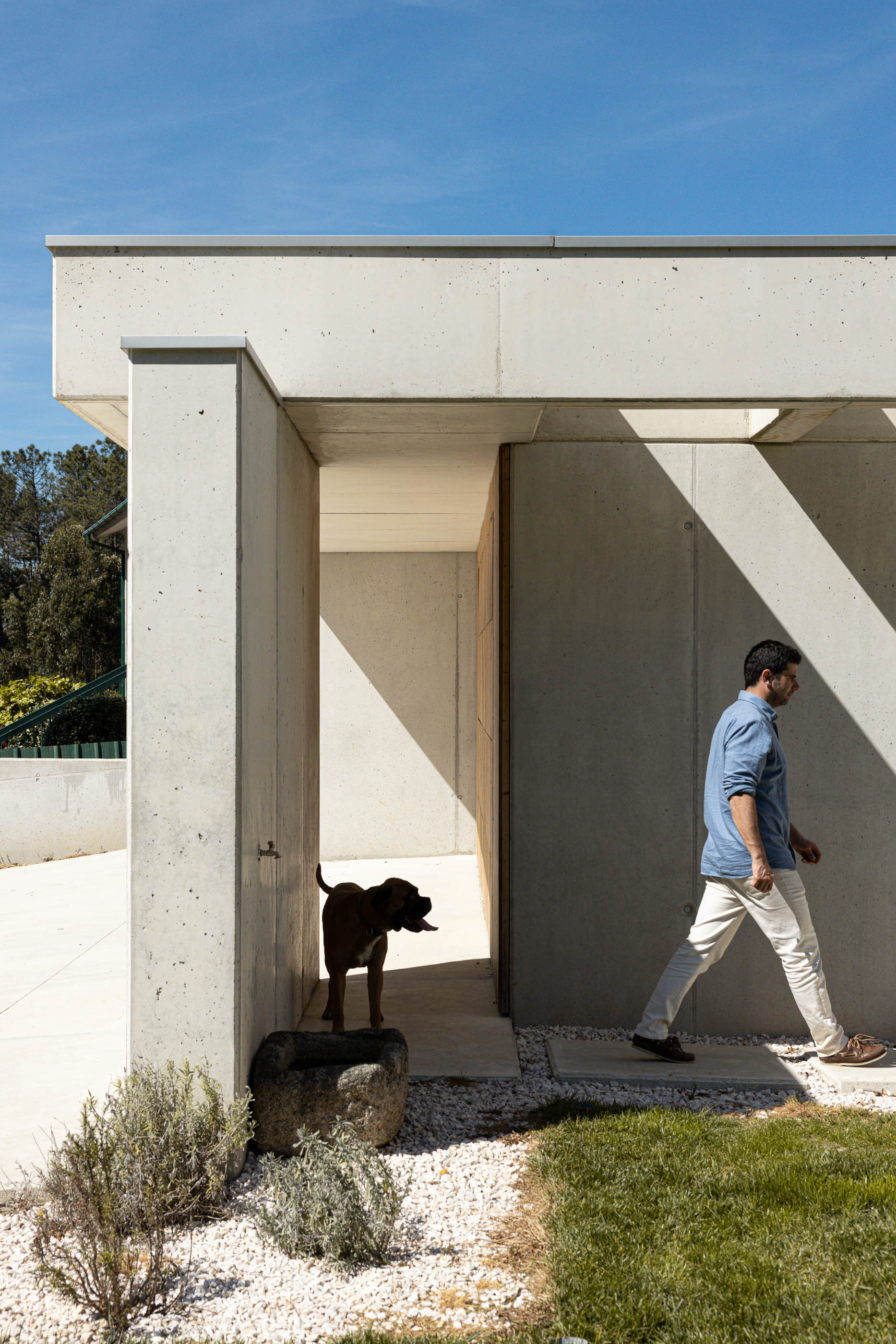 Entrada: Alpendre em betão com homem a passar e cão a beber água num bebedouro de pedra. — Casa em Mundão arquitetura Um para Um, arquitectura e engenharia em Viseu