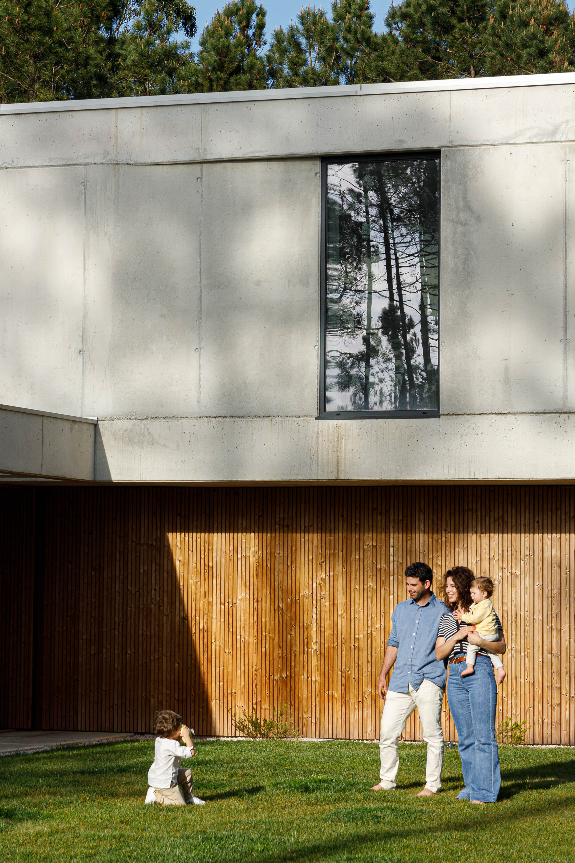Exterior: Fachada da casa com família no relvado e detalhes em betão e madeira. — Casa em Mundão arquitetura Um para Um, arquitectura e engenharia em Viseu
