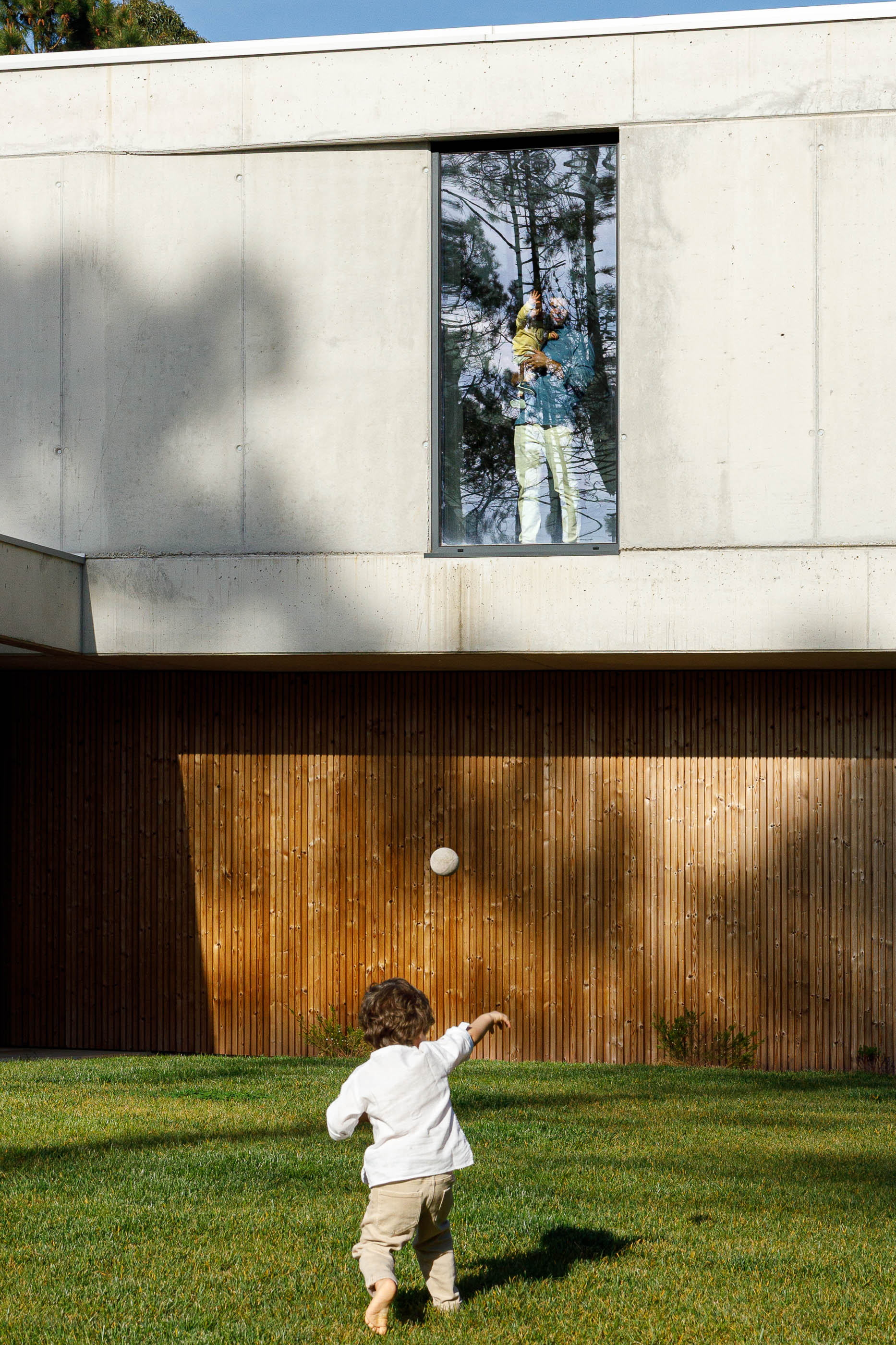 Exterior: Fachada da casa com criança a brincar no relvado e detalhes em betão e madeira. — Casa em Mundão arquitetura Um para Um, arquitectura e engenharia em Viseu
