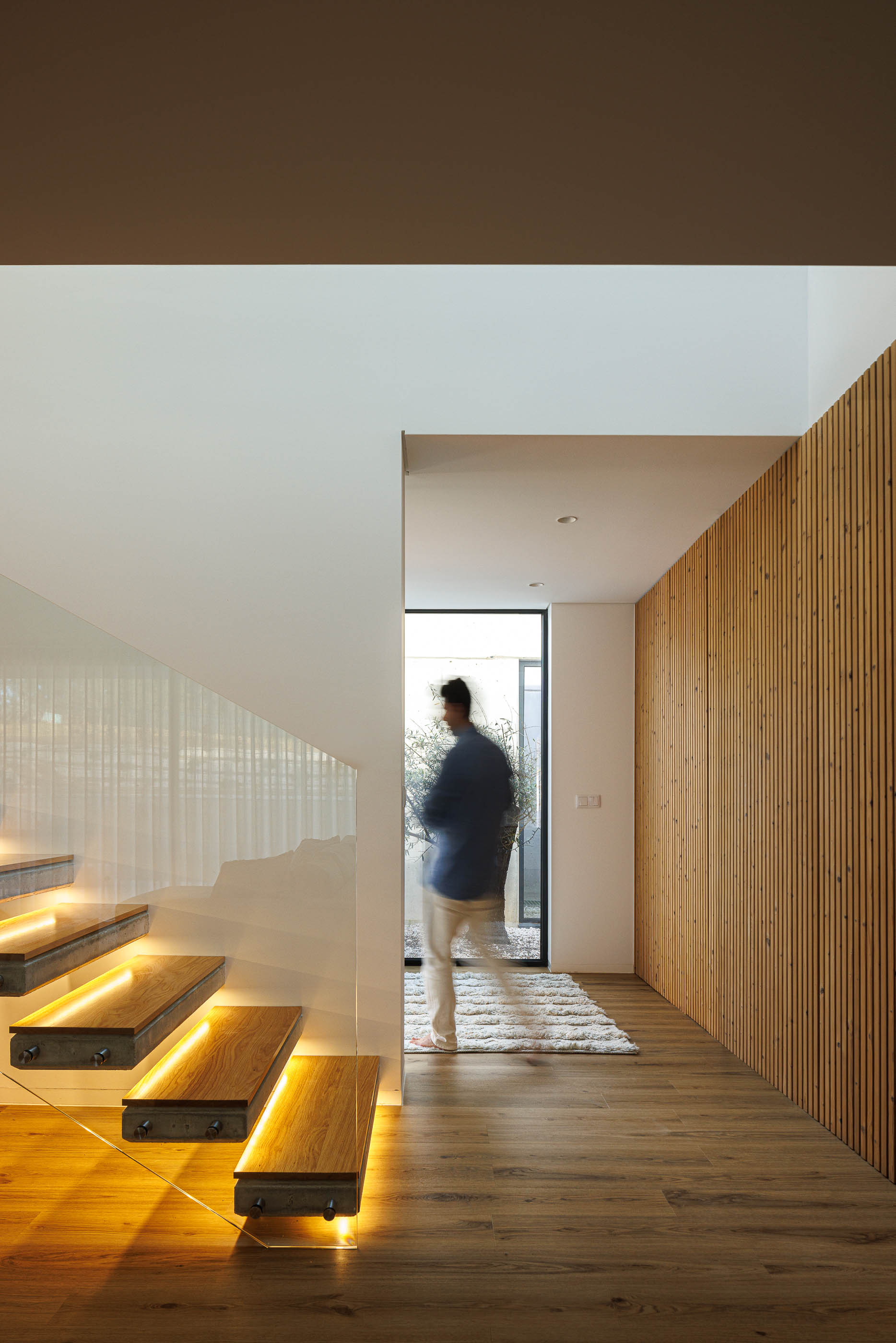 Hall: Escadas iluminadas com madeira, parede em madeira e pessoa a passar. — Casa em Mundão arquitetura Um para Um, arquitectura e engenharia em Viseu