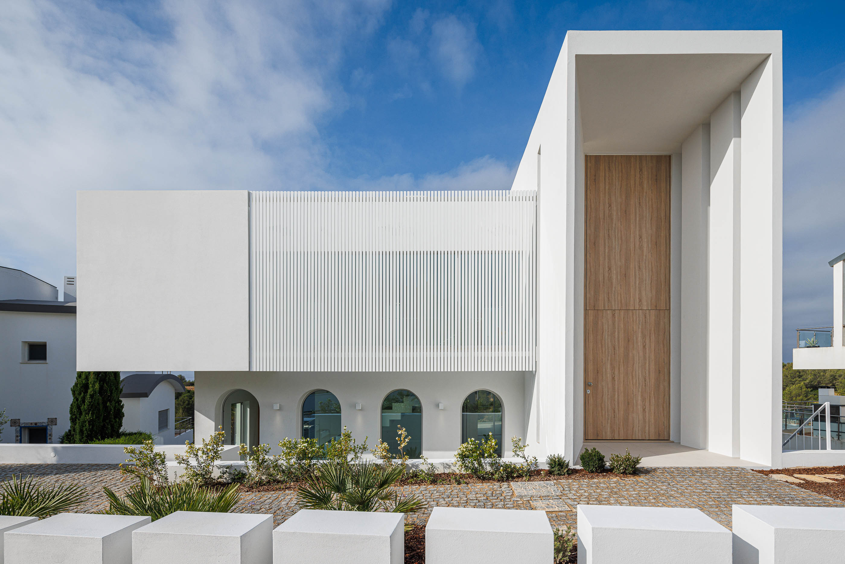 Entrada: Fachada minimalista branca com porta alta em madeira e elementos verticais. — Villa Fortaleza arquitetura Vieira de Moura Architects em Algarve