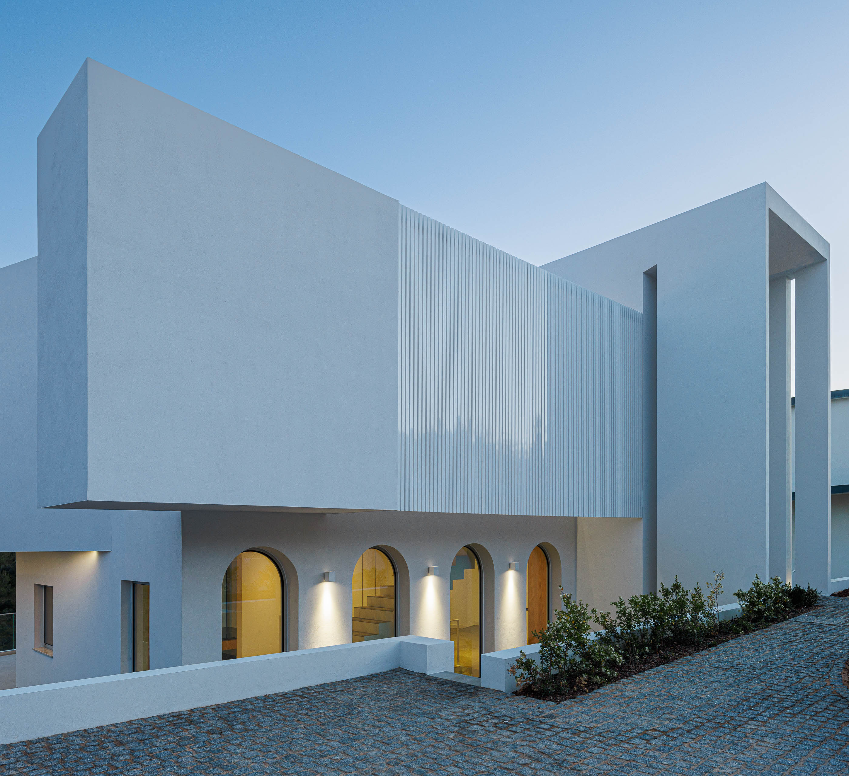 Exterior: Fachada da moradia iluminada, com arcos e porta em madeira. — Villa Fortaleza arquitetura Vieira de Moura Architects em Algarve
