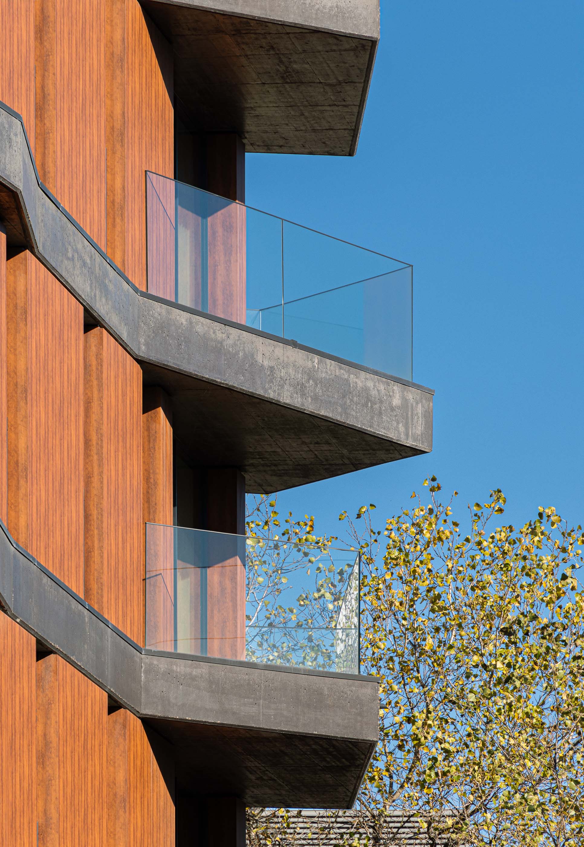 Fachada: Detalhe das varandas em betão com guardas de vidro, com revestimento exterior em madeira e céu azul. — Puro Tower arquitetura Mário Alves Arquitecto em Aveiro