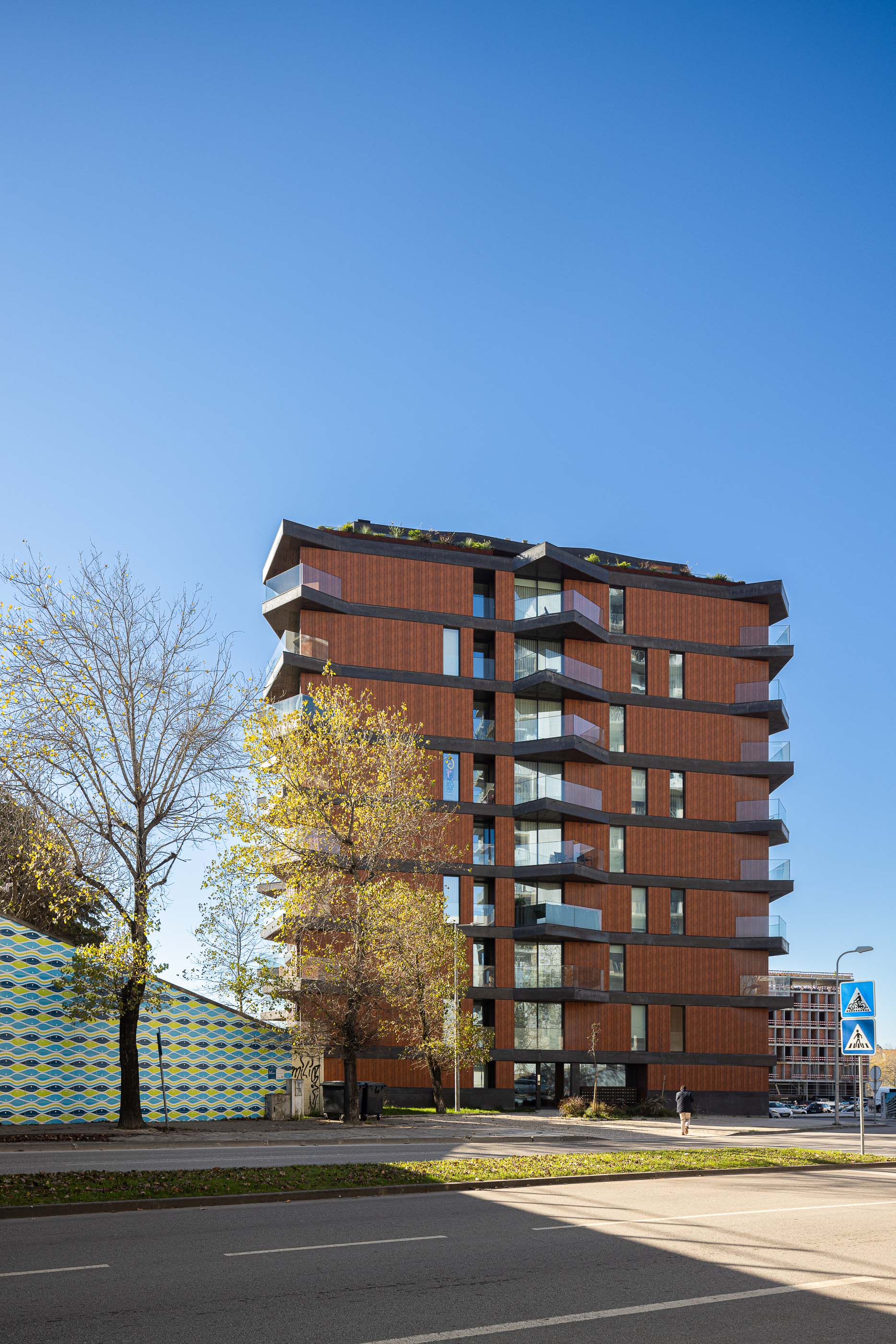 Fachada: Vista frontal da torre residencial com varandas triangulares e revestimento em madeira, junto à rua. — Puro Tower arquitetura Mário Alves Arquitecto em Aveiro