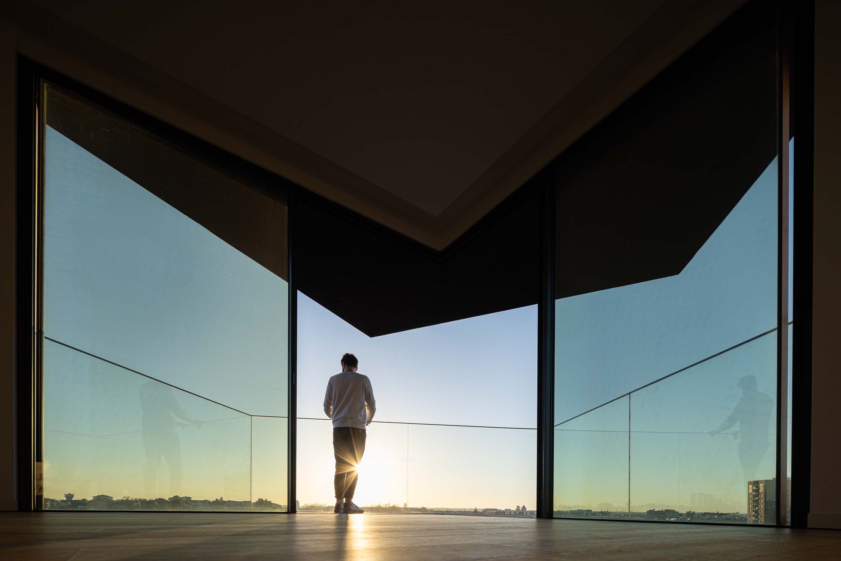 Varanda: Homem a olhar para a paisagem numa varanda com grandes janelas envidraçadas e pavimento em madeira. — Puro Tower arquitetura Mário Alves Arquitecto em Aveiro