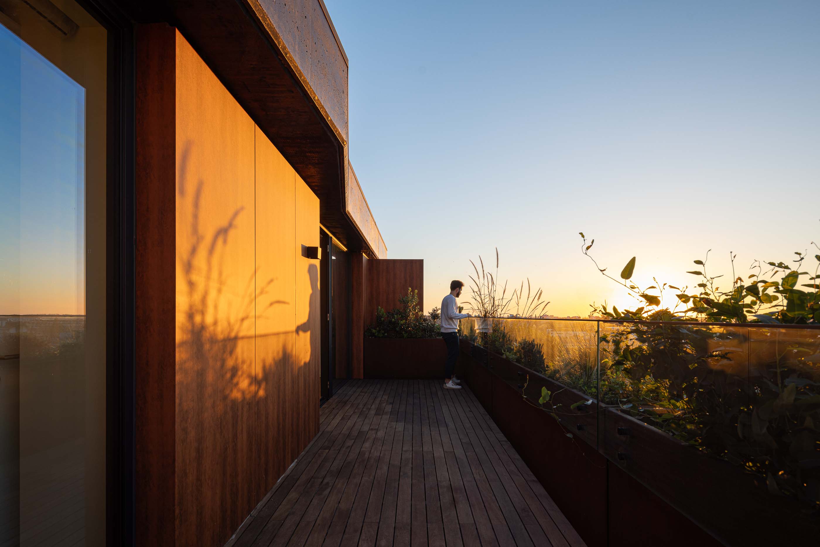 Varanda: Homem numa varanda com pavimento em madeira e vegetação, com vista para a cidade ao pôr do sol. — Puro Tower arquitetura Mário Alves Arquitecto em Aveiro