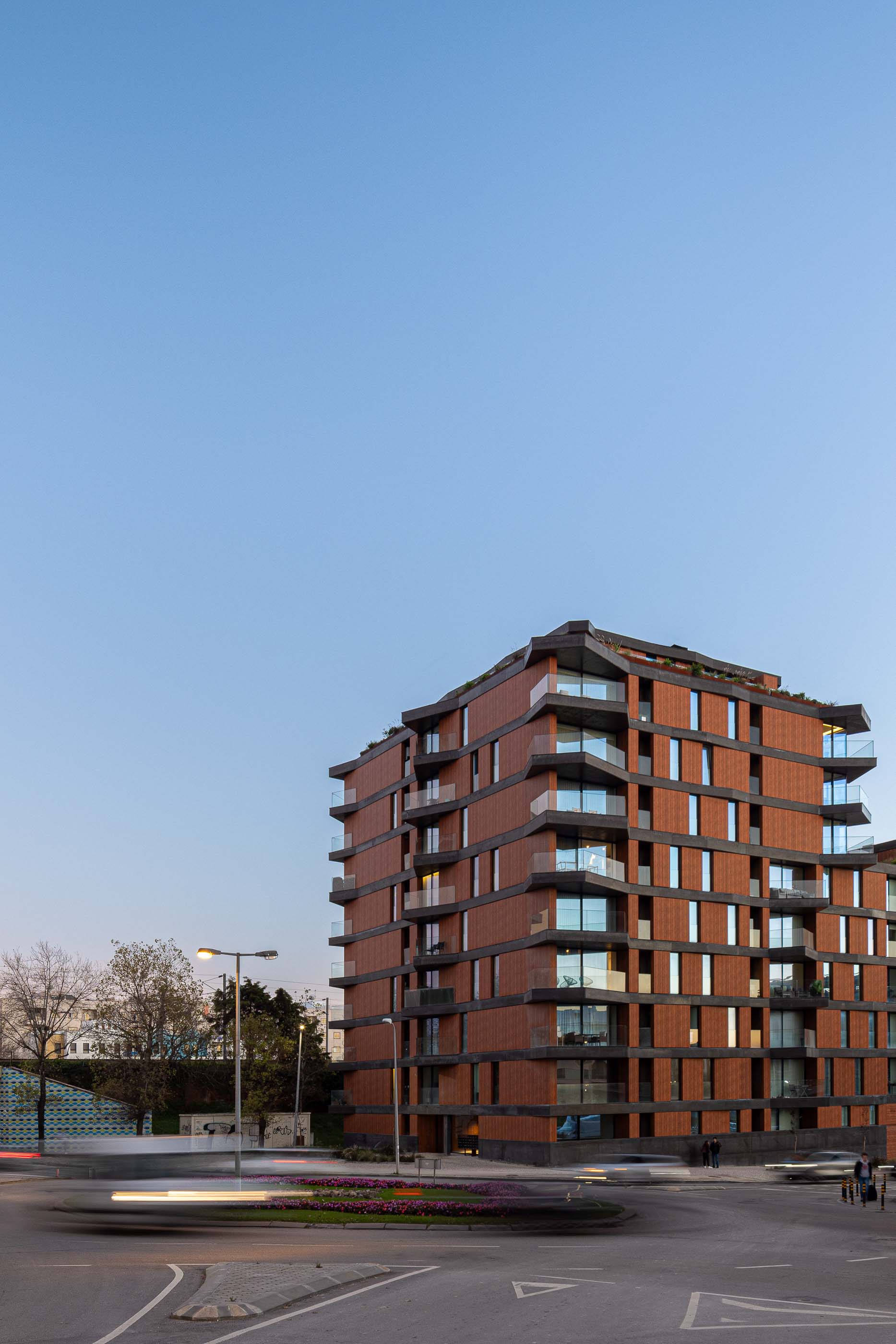 Exterior: Vista noturna da torre residencial com varandas e revestimento em madeira, junto a uma rotunda. — Puro Tower arquitetura Mário Alves Arquitecto em Aveiro