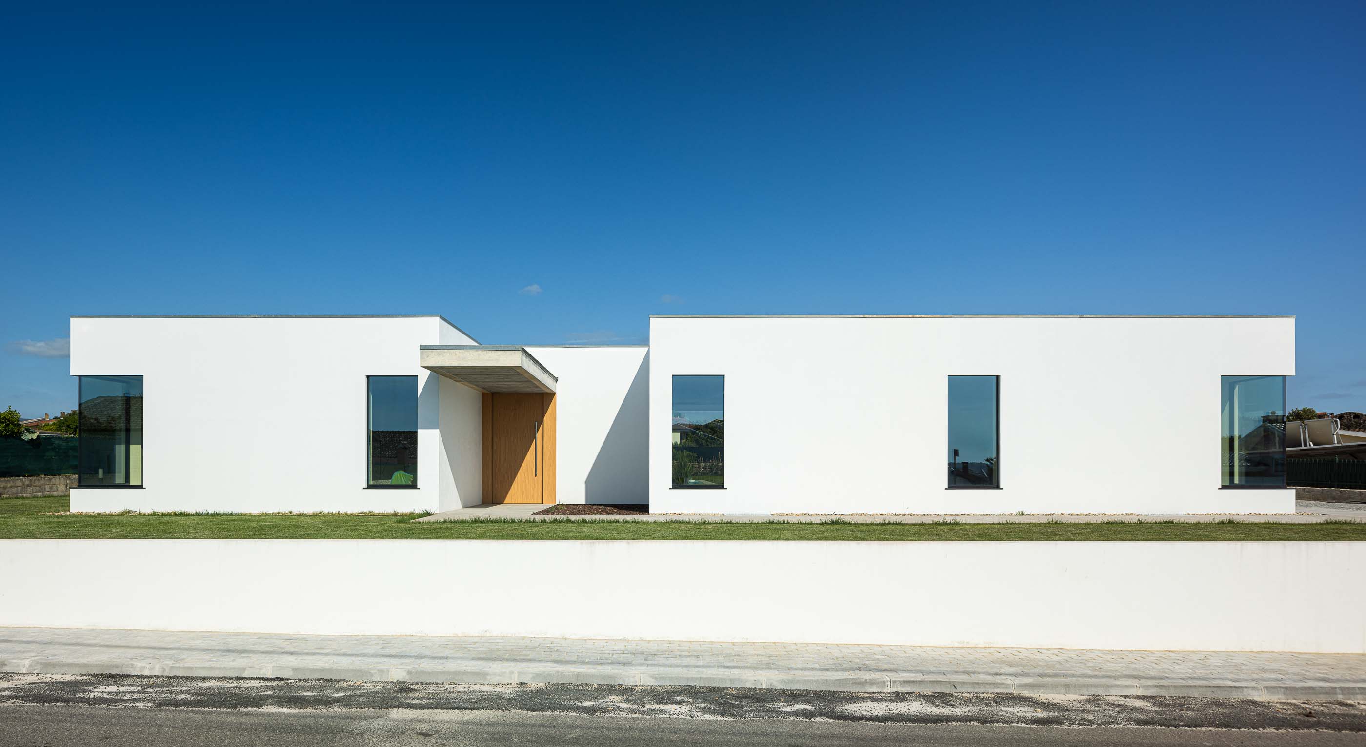 Exterior: Fachada da moradia térrea branca com porta de madeira e alpendre de betão. — Casa CS arquitetura Jorge Gonzalez, WALAA em Águeda