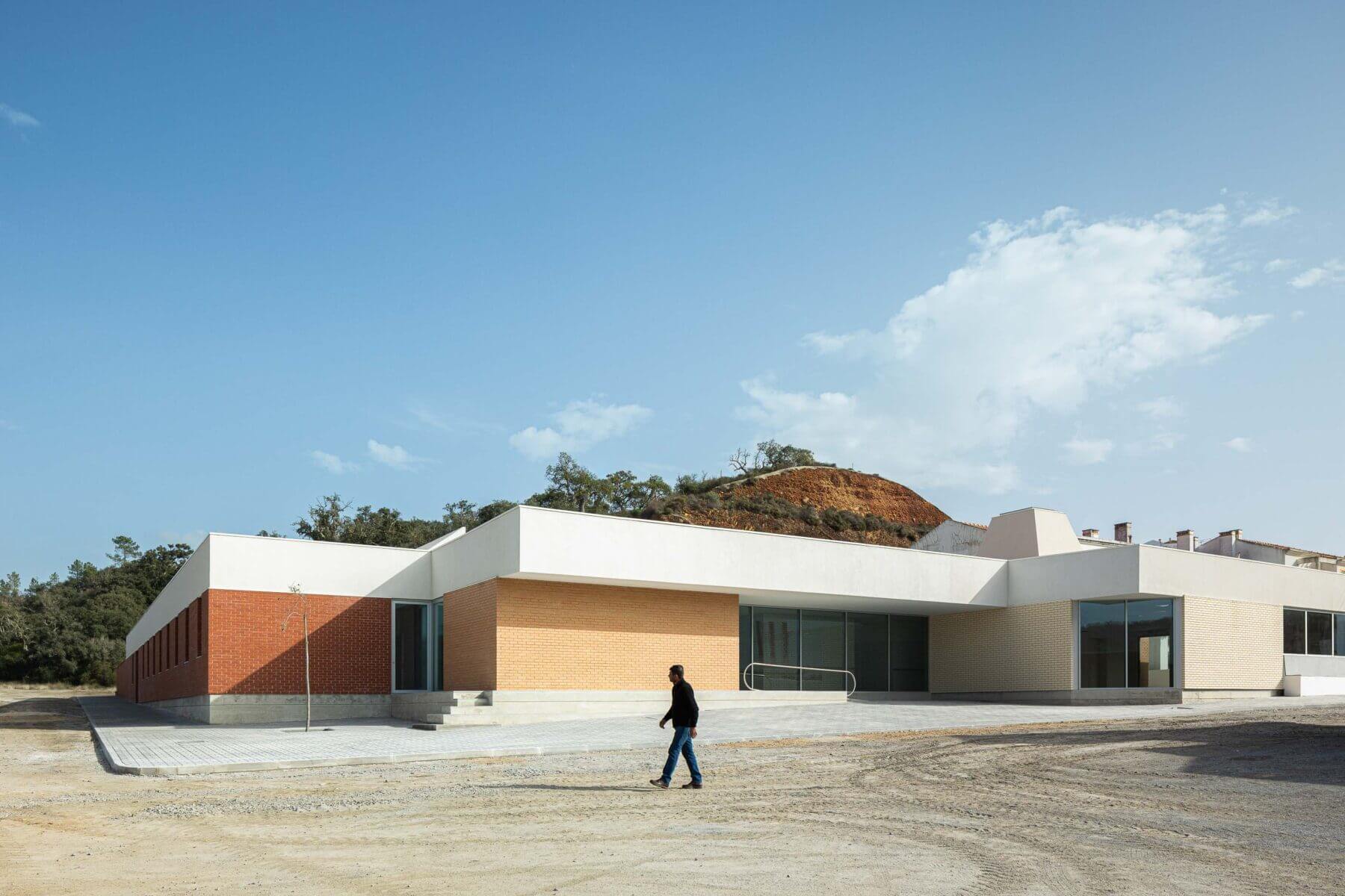 Edifício: Fachada principal da APCO, com revestimento em tijolo claro e escuro, e um homem a caminhar em frente. — Associação Paralisia Cerebral de Odemira – APCO arquitetura Filipe Xavier Oliveira em Odemira