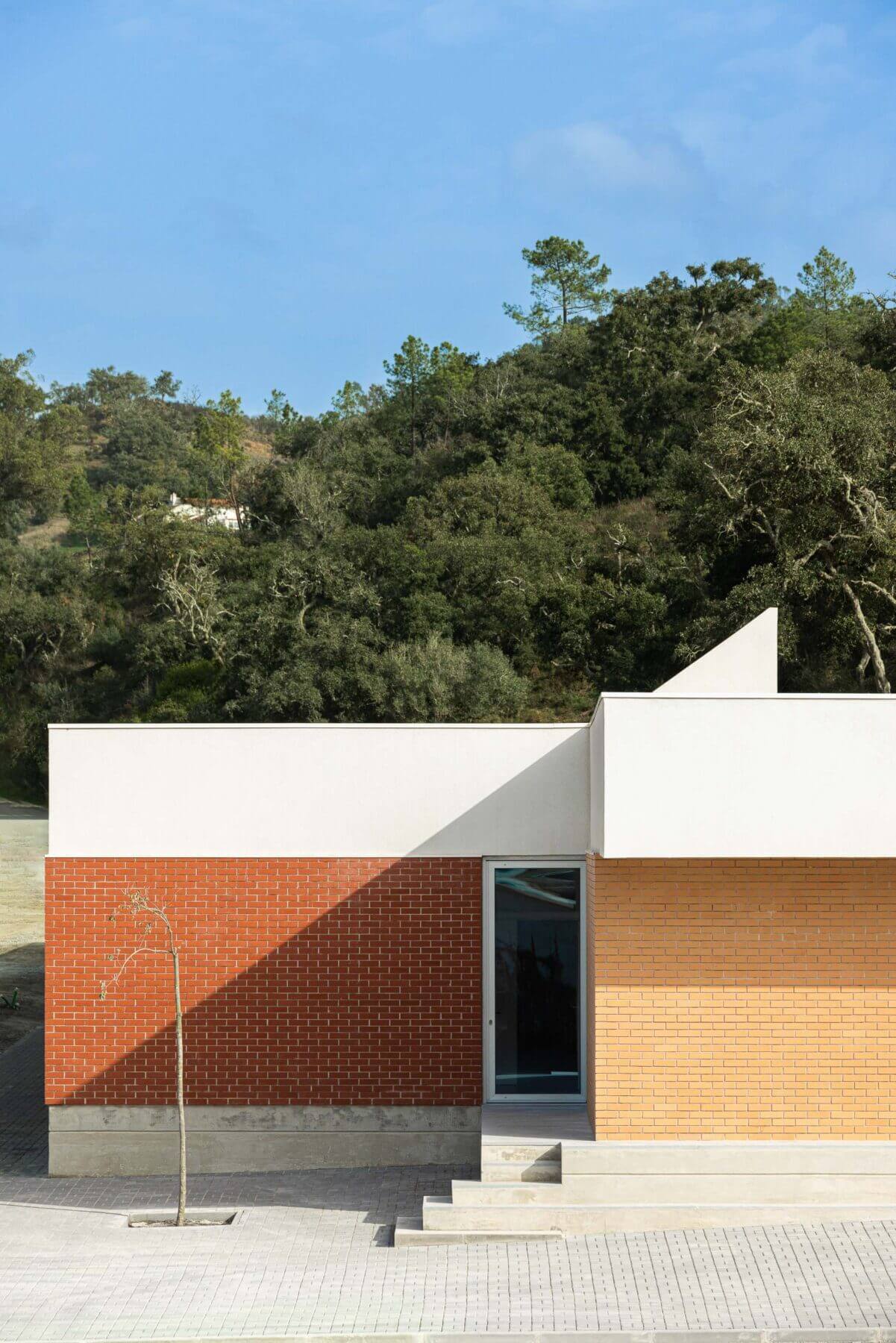 Edifício: Detalhe da fachada da APCO, com tijolo em dois tons, porta envidraçada e vegetação ao fundo. — Associação Paralisia Cerebral de Odemira – APCO arquitetura Filipe Xavier Oliveira em Odemira