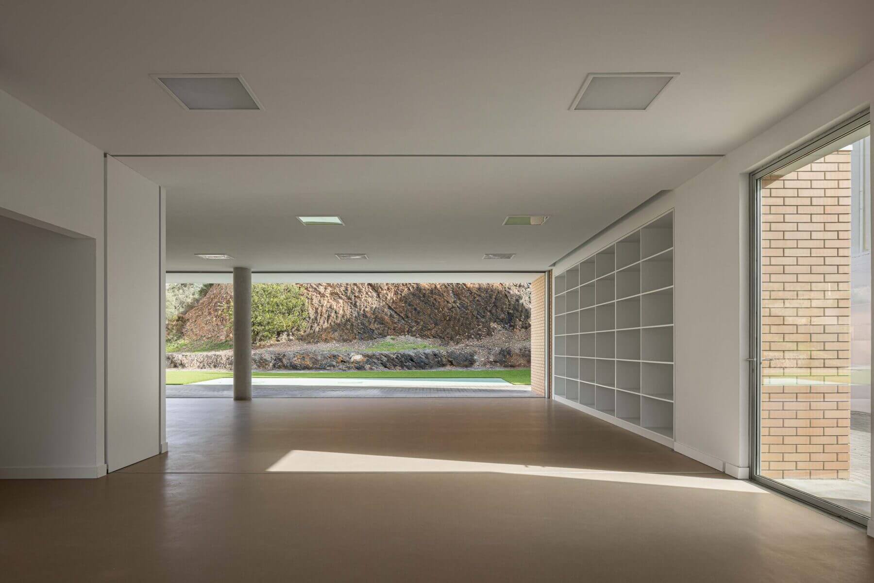 Sala polivalente: Interior da sala com pavimento castanho, estantes e vista para o exterior com relvado e muro de pedra. — Associação Paralisia Cerebral de Odemira – APCO arquitetura Filipe Xavier Oliveira em Odemira