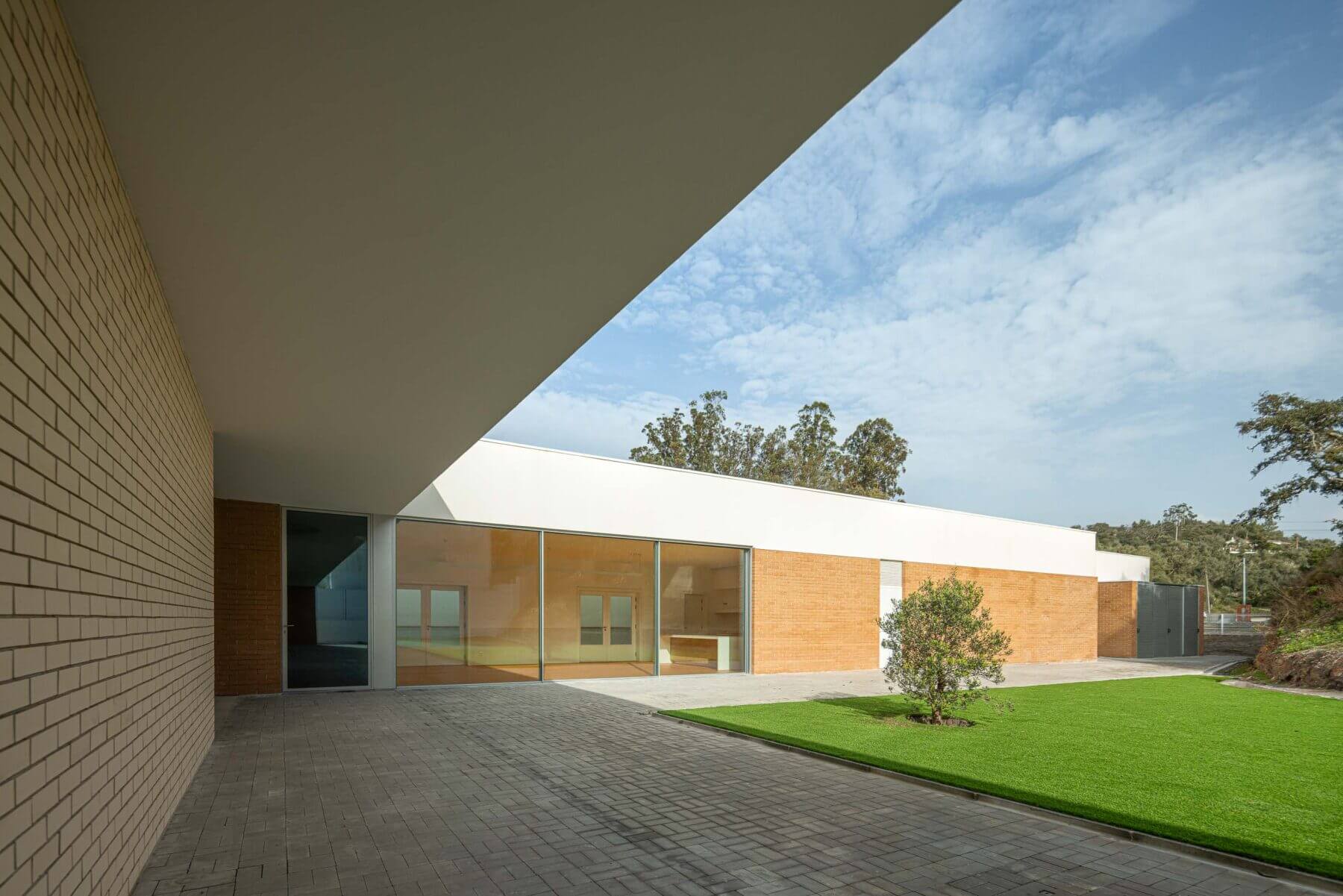 Pátio: Vista do pátio exterior com relvado, pavimento em calçada e fachada em tijolo da APCO. — Associação Paralisia Cerebral de Odemira – APCO arquitetura Filipe Xavier Oliveira em Odemira