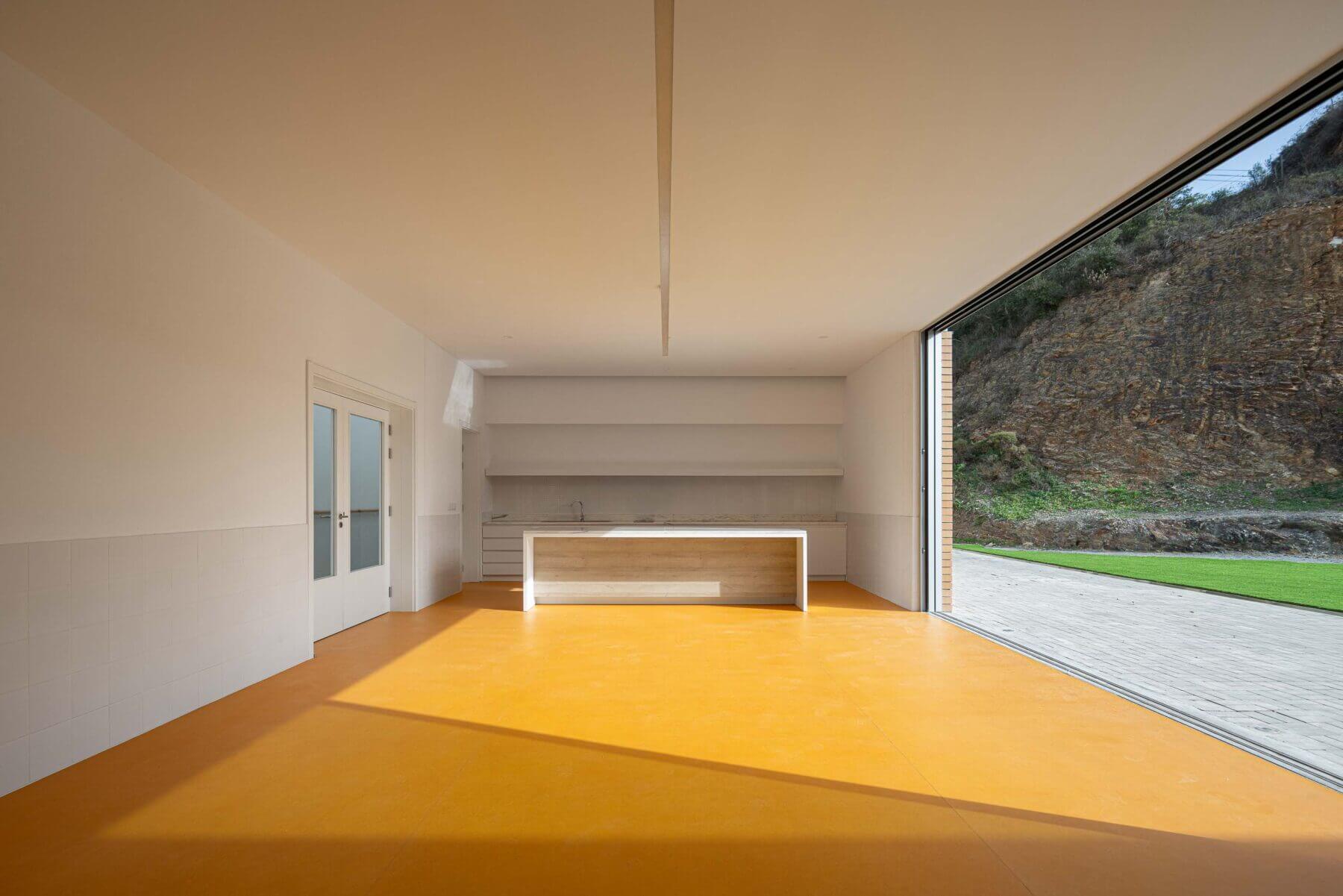 Cozinha: Pavimento amarelo, bancada central em madeira clara e grandes portas de vidro deslizantes para o exterior. — Associação Paralisia Cerebral de Odemira – APCO arquitetura Filipe Xavier Oliveira em Odemira