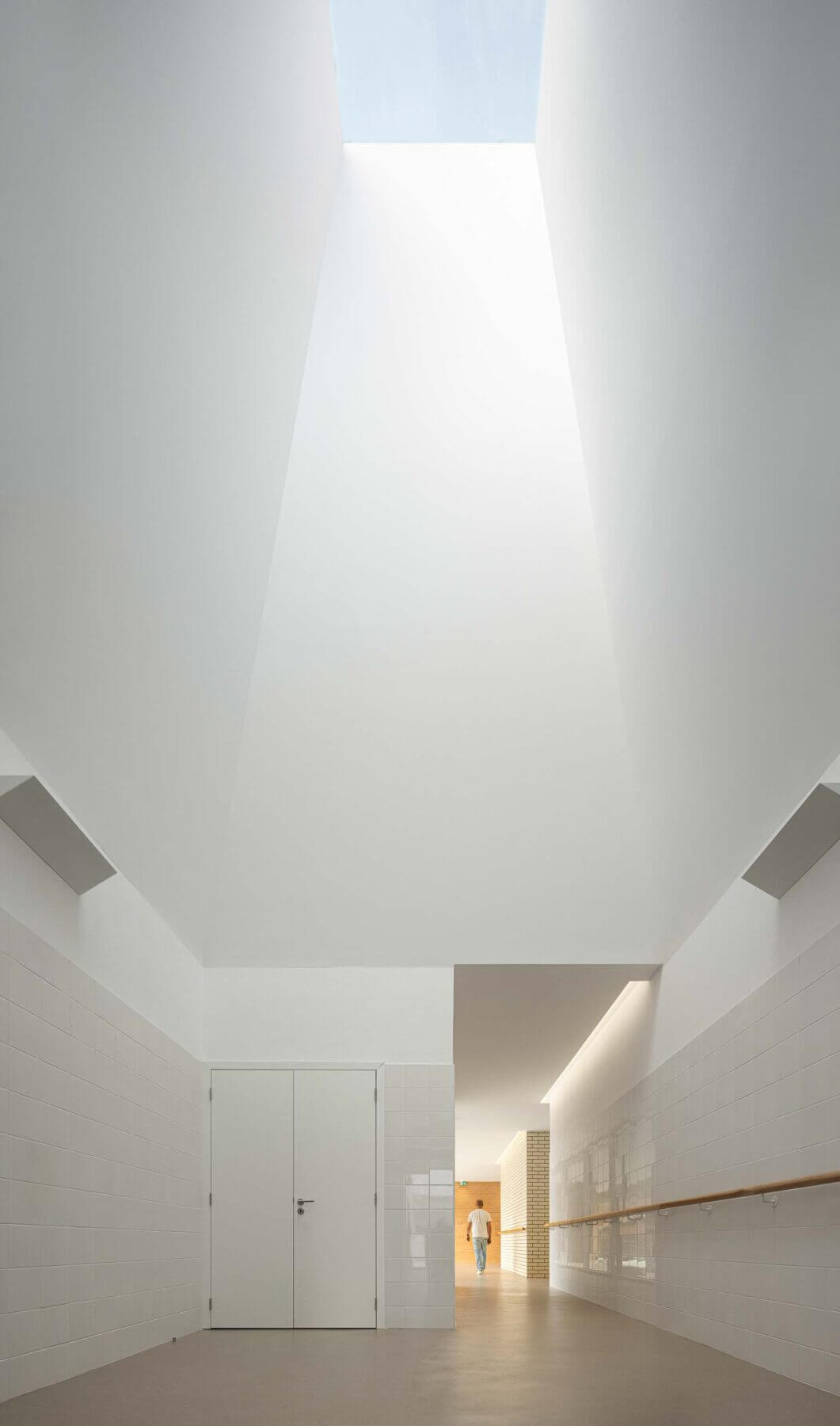 Corredor: Perspectiva do corredor com luz zenital, pavimento castanho, azulejos brancos e portas laterais. — Associação Paralisia Cerebral de Odemira – APCO arquitetura Filipe Xavier Oliveira em Odemira