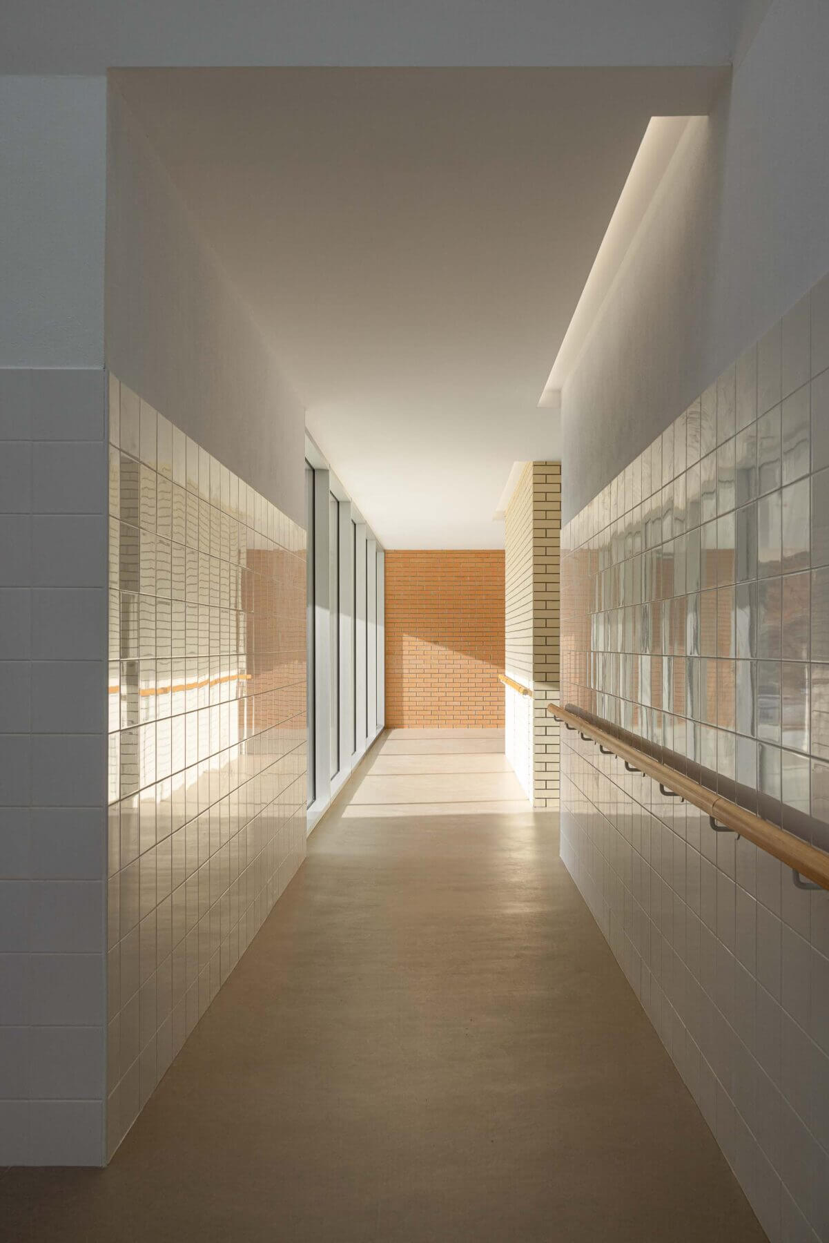 Corredor: Corredor com azulejos brancos, corrimão de madeira, pavimento castanho e luz natural. — Associação Paralisia Cerebral de Odemira – APCO arquitetura Filipe Xavier Oliveira em Odemira