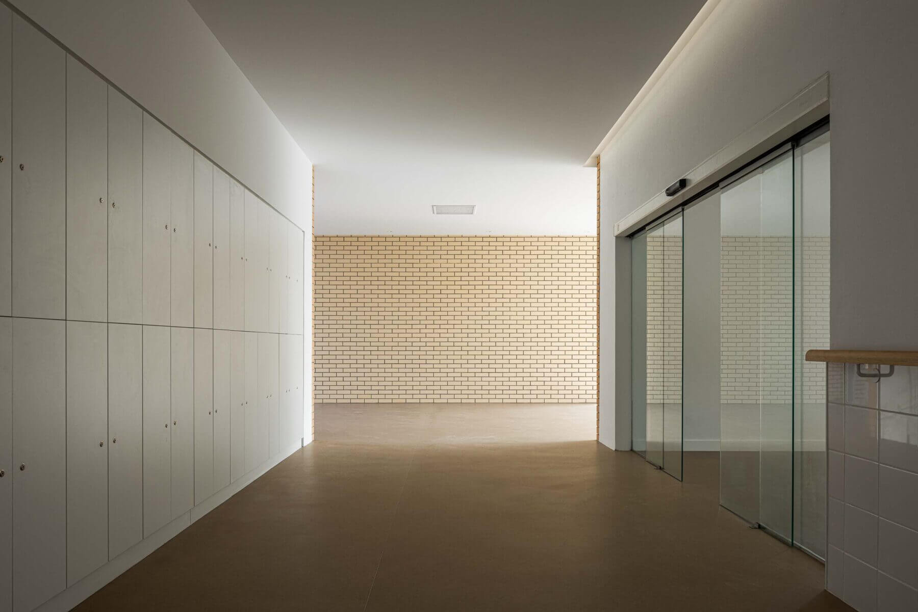 Corredor: Corredor com cacifos brancos, pavimento castanho e porta de vidro deslizante. — Associação Paralisia Cerebral de Odemira – APCO arquitetura Filipe Xavier Oliveira em Odemira