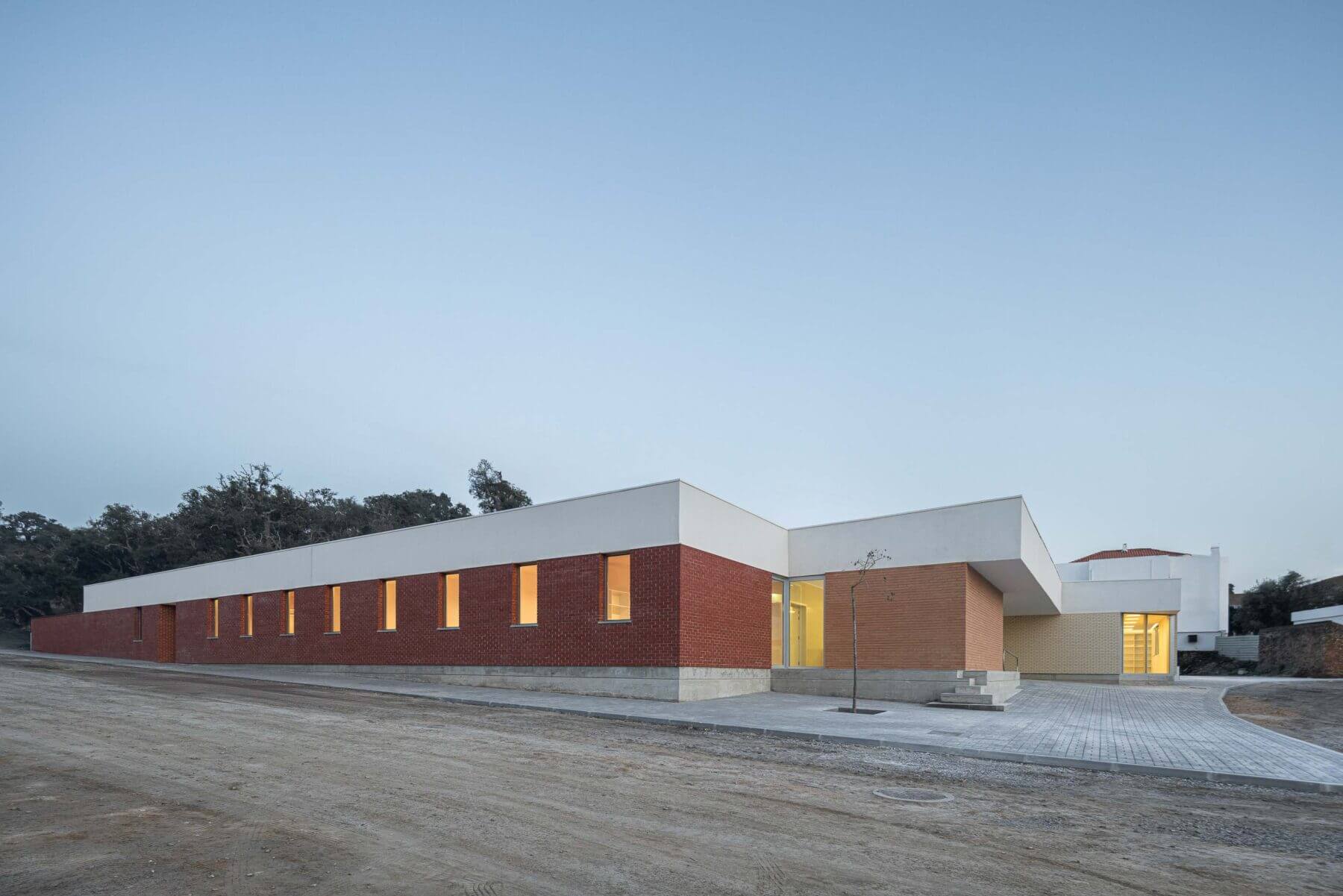 Edifício: Vista noturna da fachada da APCO, com iluminação interior e exterior, destacando o tijolo e o branco. — Associação Paralisia Cerebral de Odemira – APCO arquitetura Filipe Xavier Oliveira em Odemira