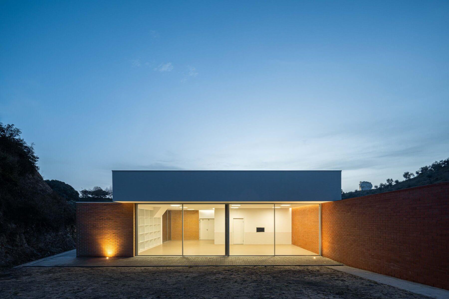 Sala polivalente: Fachada envidraçada iluminada da sala polivalente, com estantes brancas e paredes em tijolo. — Associação Paralisia Cerebral de Odemira – APCO arquitetura Filipe Xavier Oliveira em Odemira