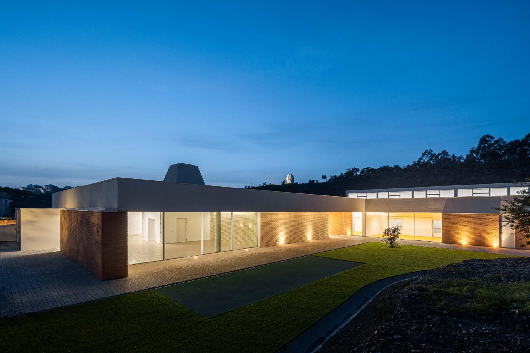 Edifício: Vista noturna da APCO com relvado, fachada em tijolo e iluminação exterior. — Associação Paralisia Cerebral de Odemira – APCO arquitetura Filipe Xavier Oliveira em Odemira