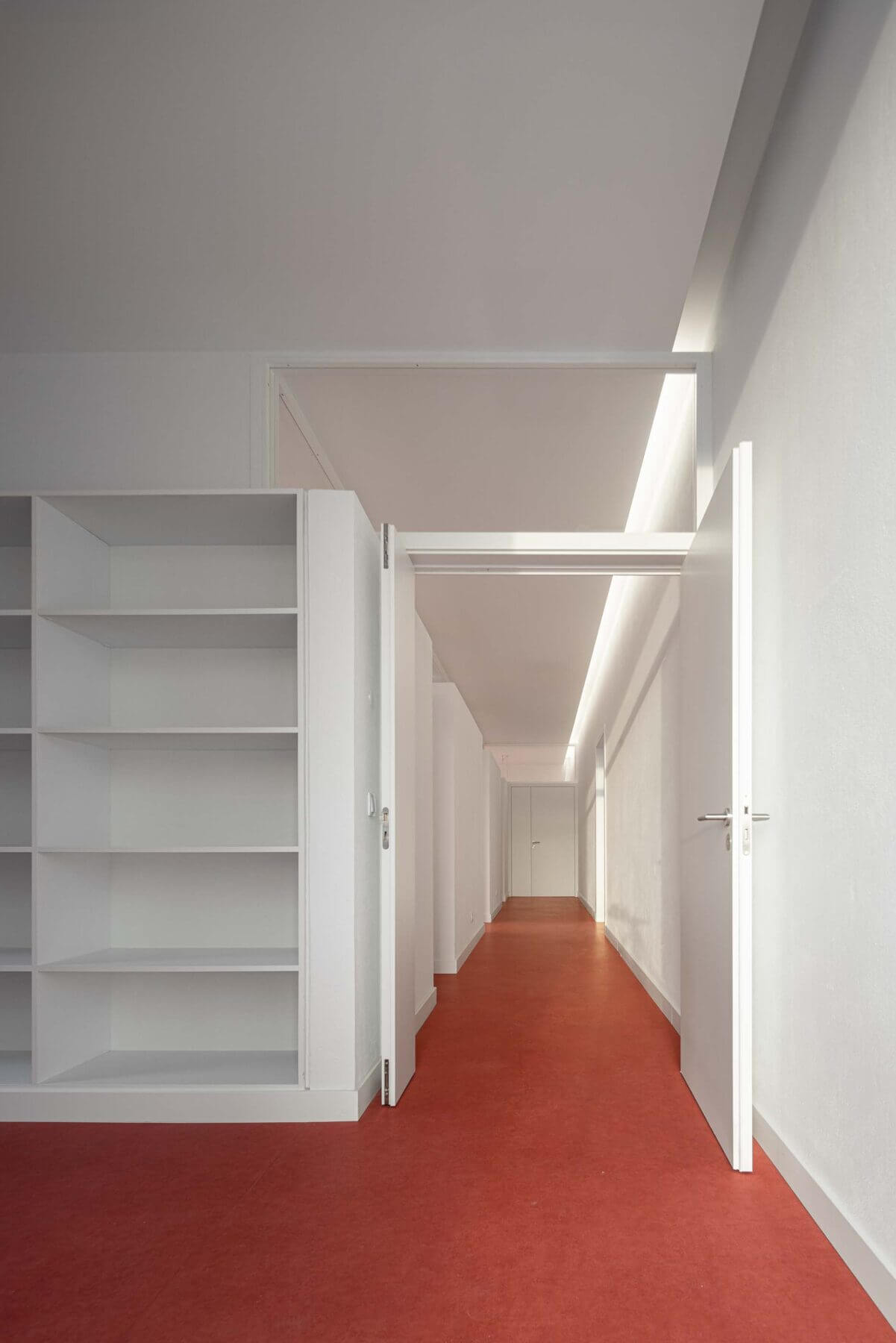Sala de atividades: Sala com estantes brancas, pavimento vermelho e porta aberta para o corredor iluminado. — Associação Paralisia Cerebral de Odemira – APCO arquitetura Filipe Xavier Oliveira em Odemira