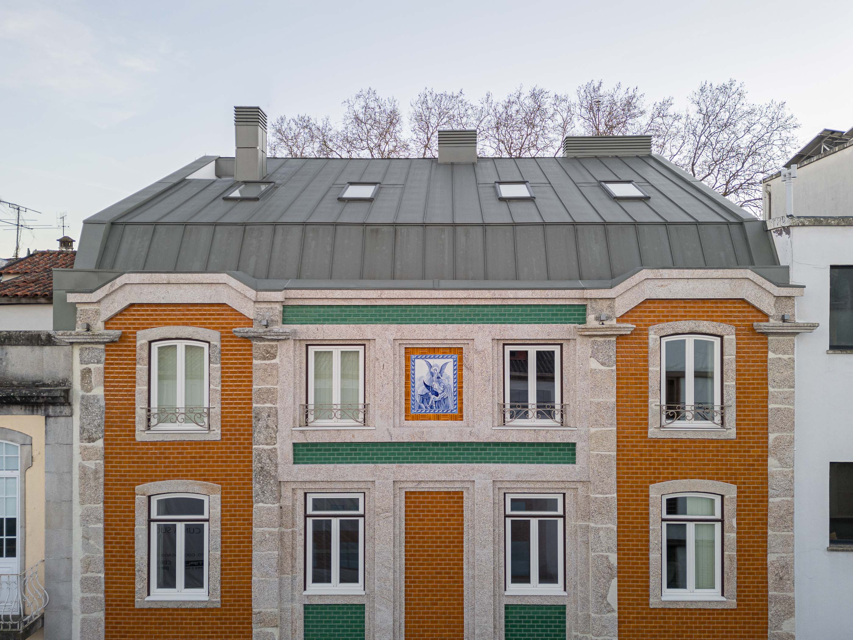Fachada: Edifício com tijoleira laranja, granito e painel de azulejos, sob telhado cinzento com janelas de sótão. — Apartamentos Vitória arquitetura Floret Arquitectura em Viseu