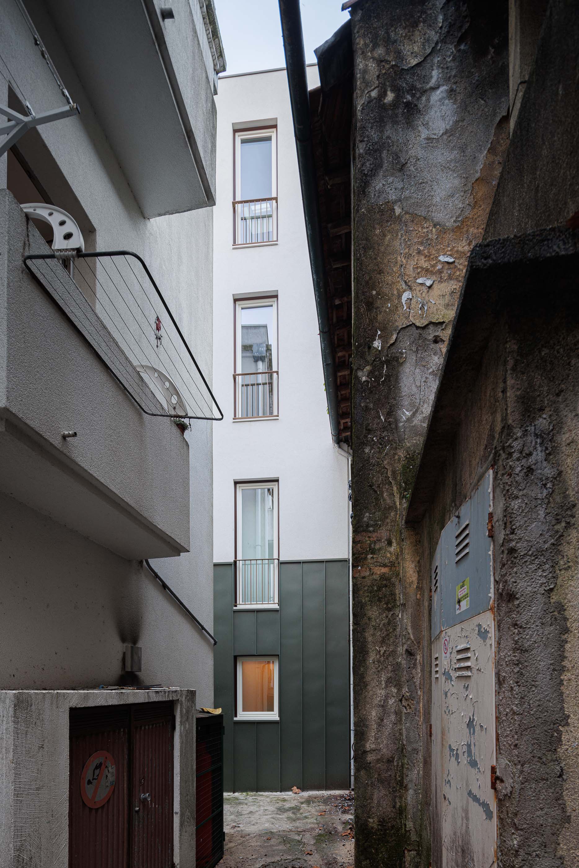 Exterior: Vista estreita da fachada lateral do edifício entre prédios antigos, com revestimento cinzento e janelas. — Apartamentos Vitória arquitetura Floret Arquitectura em Viseu