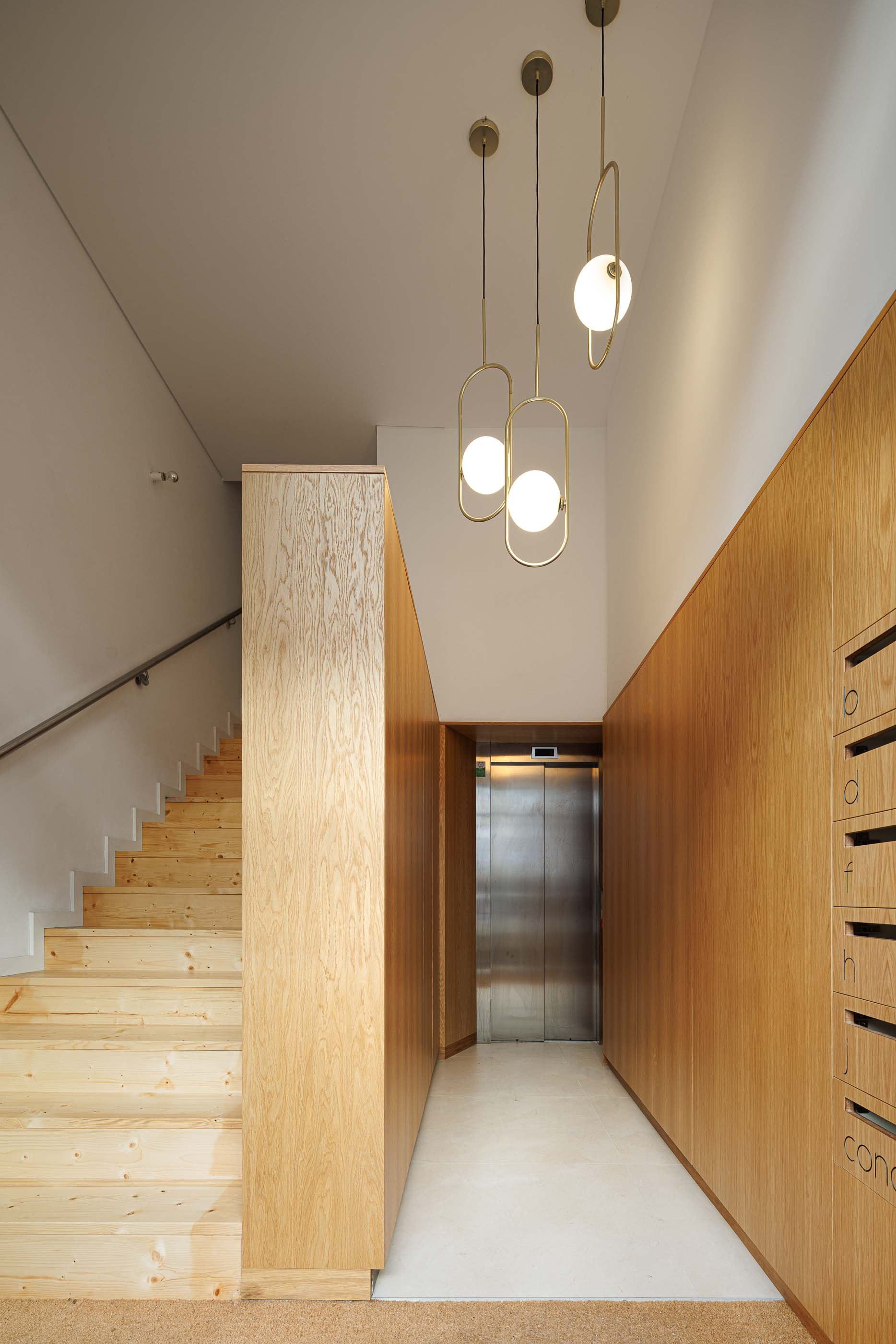 Hall de entrada: Escadas em madeira clara, elevador e parede com caixas de correio sob candeeiros suspensos. — Apartamentos Vitória arquitetura Floret Arquitectura em Viseu
