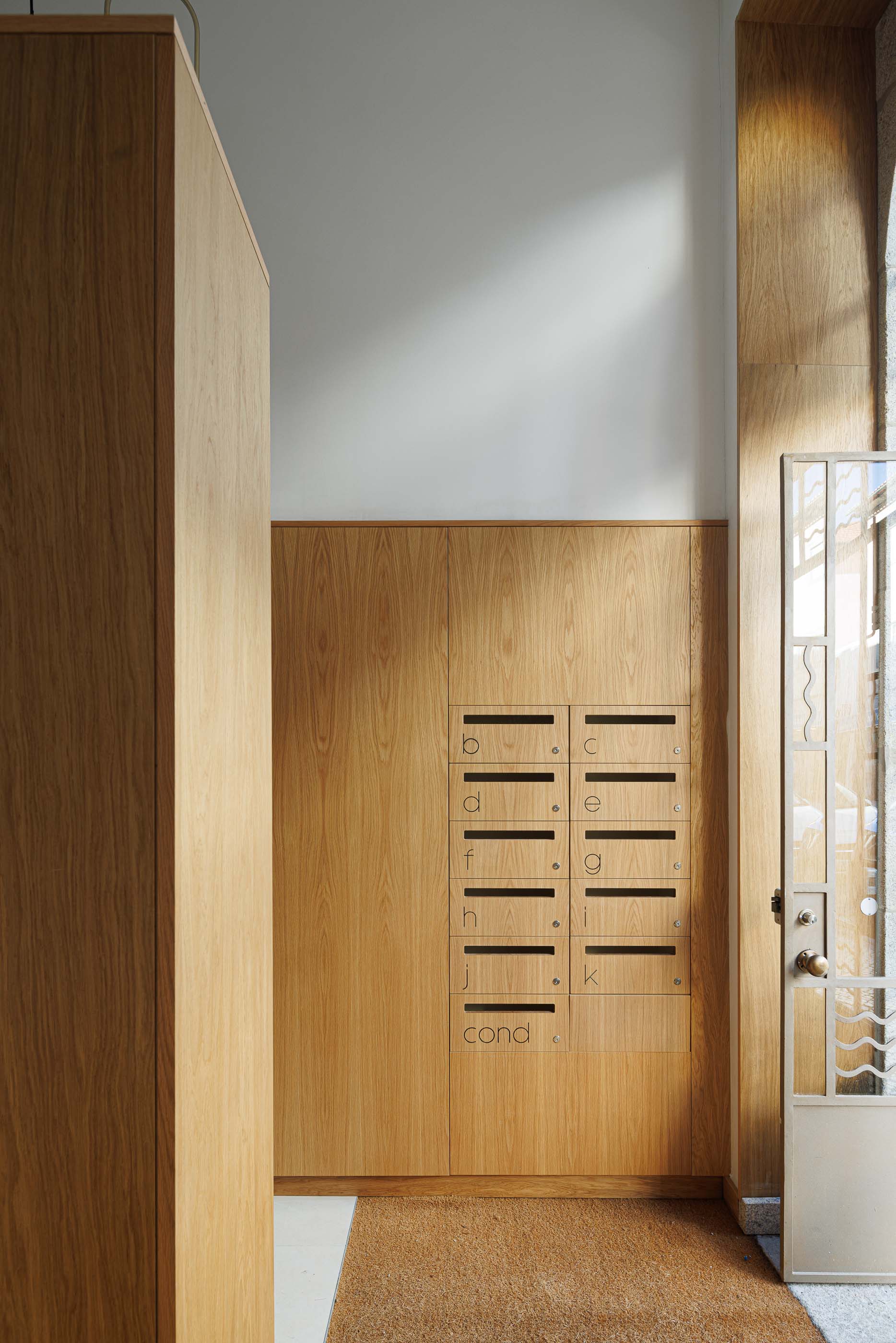 Hall de entrada: Caixas de correio em madeira clara, tapete de fibra natural e porta de entrada envidraçada. — Apartamentos Vitória arquitetura Floret Arquitectura em Viseu