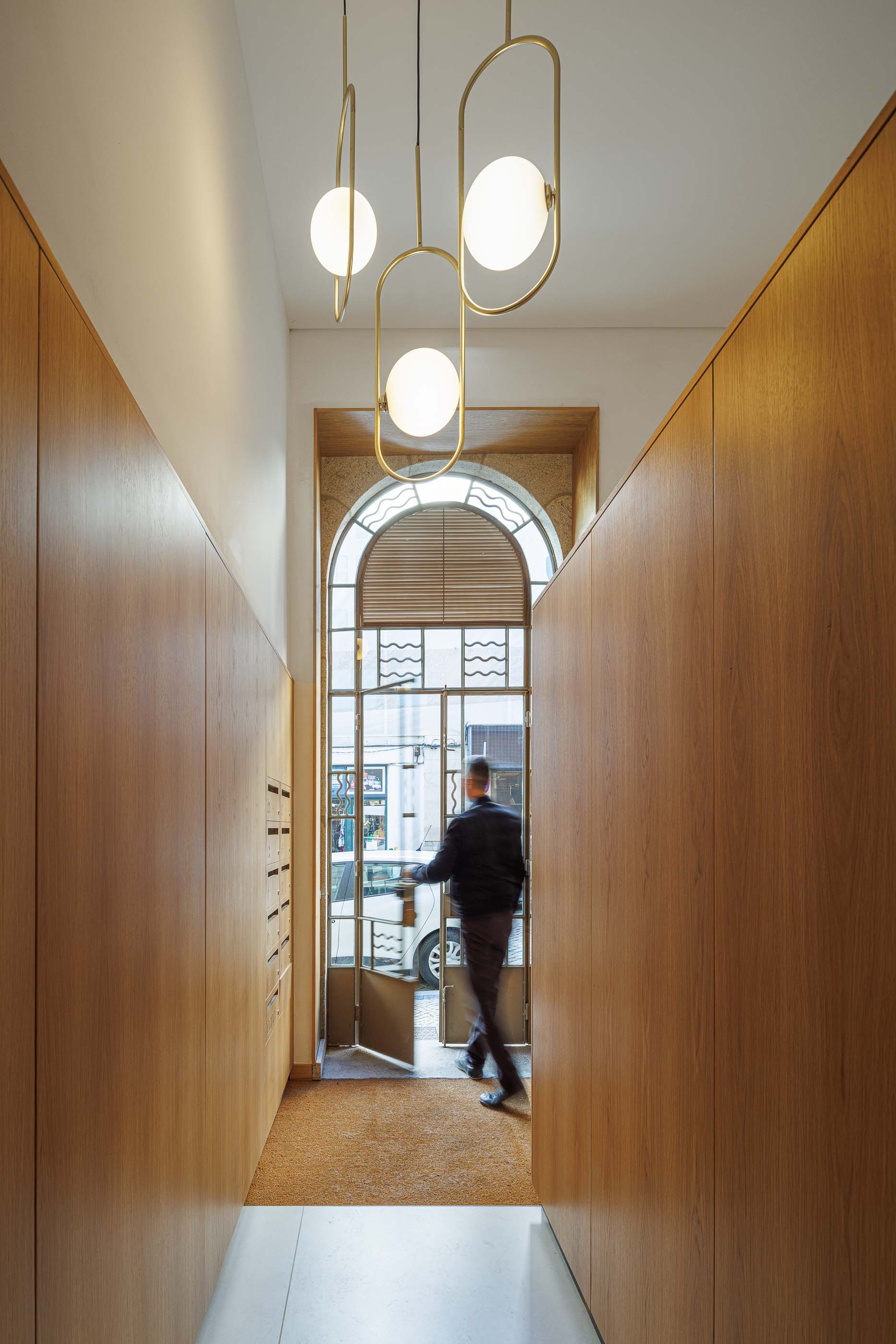 Hall de entrada: Pessoa a entrar no edifício, com caixas de correio e candeeiros suspensos. — Apartamentos Vitória arquitetura Floret Arquitectura em Viseu