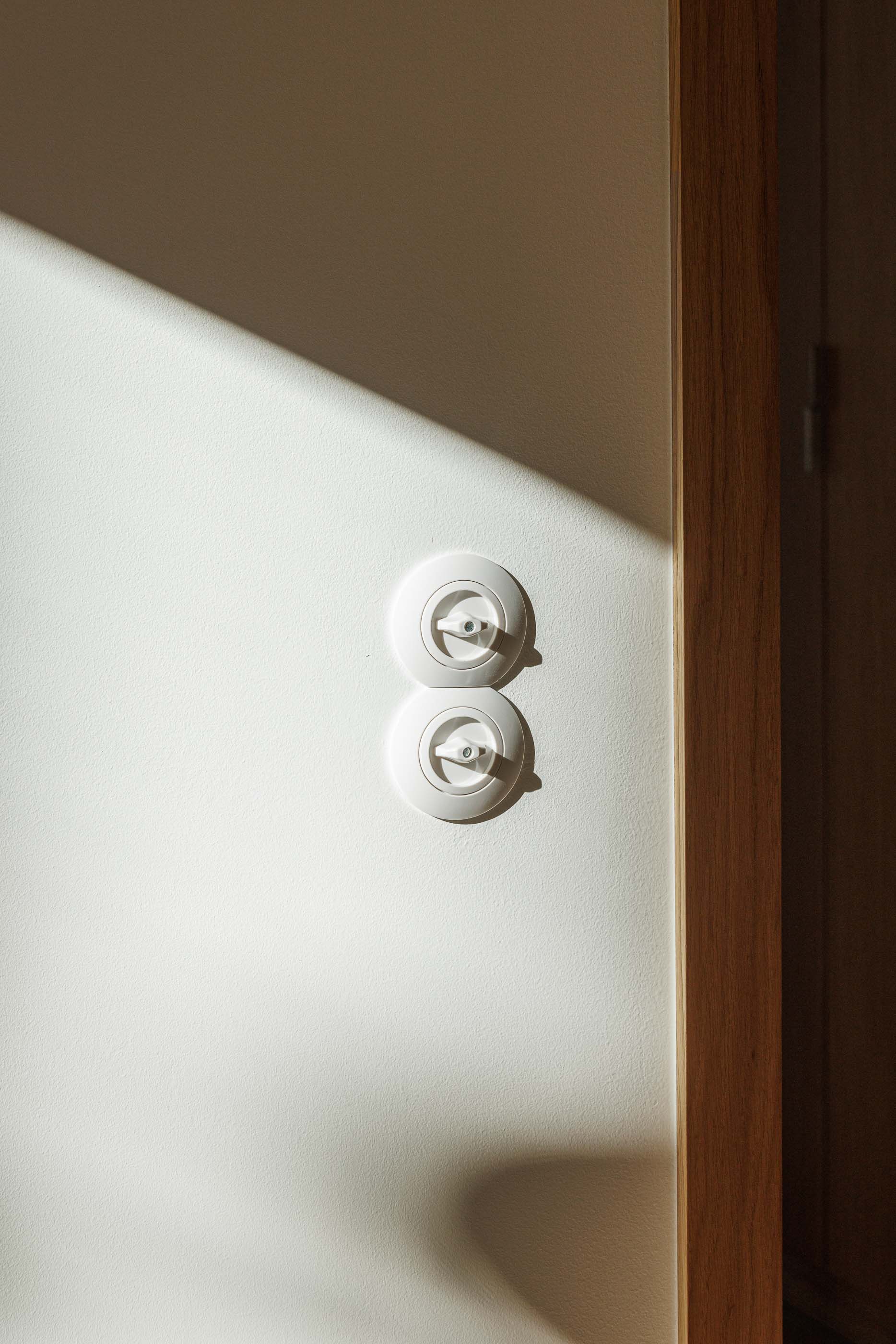 Detalhe: Interruptores rotativos brancos numa parede branca com caixilharia em madeira clara. — Apartamentos Vitória arquitetura Floret Arquitectura em Viseu