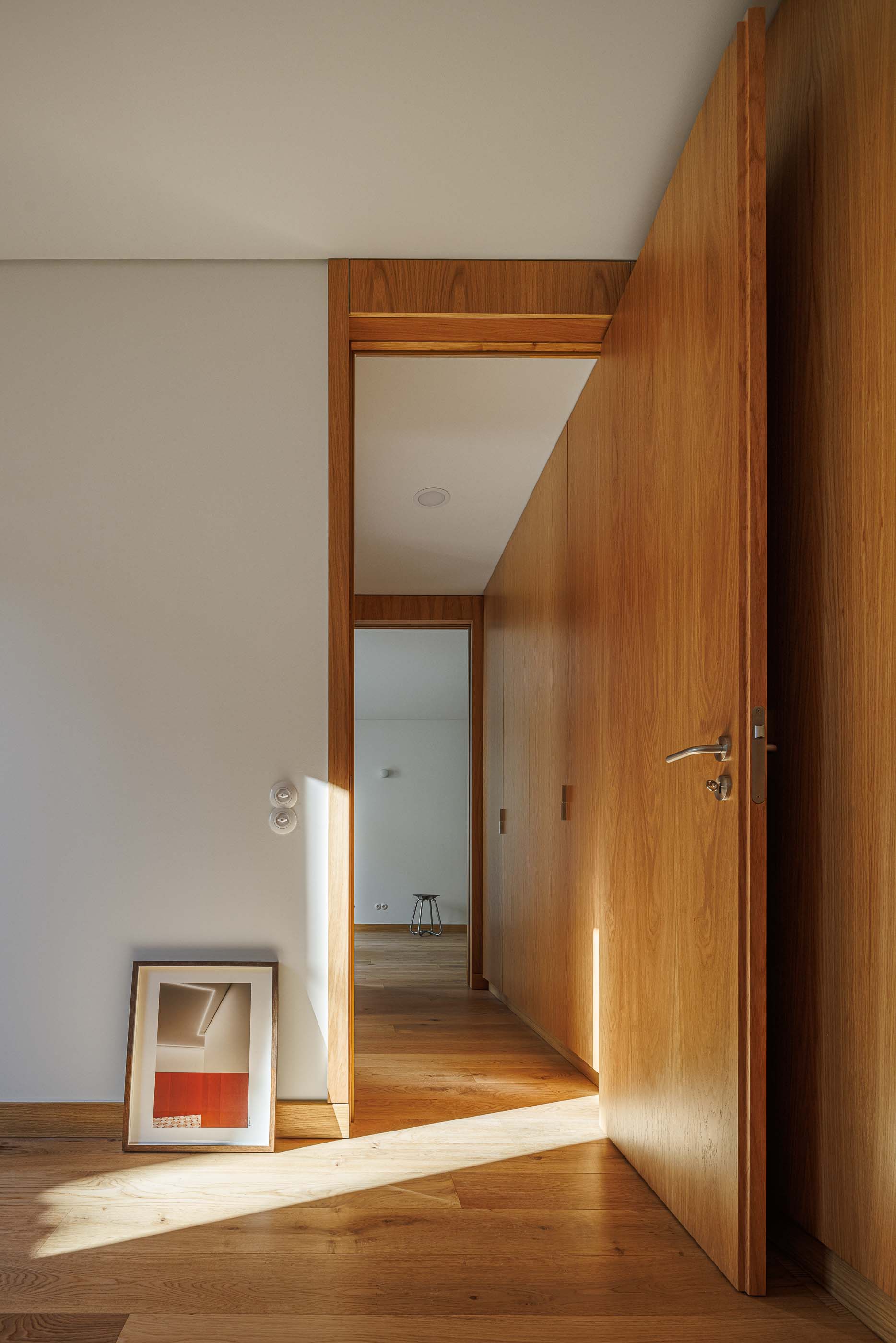 Quarto: Porta aberta para corredor com armários embutidos, pavimento em madeira e obra de arte no chão. — Apartamentos Vitória arquitetura Floret Arquitectura em Viseu