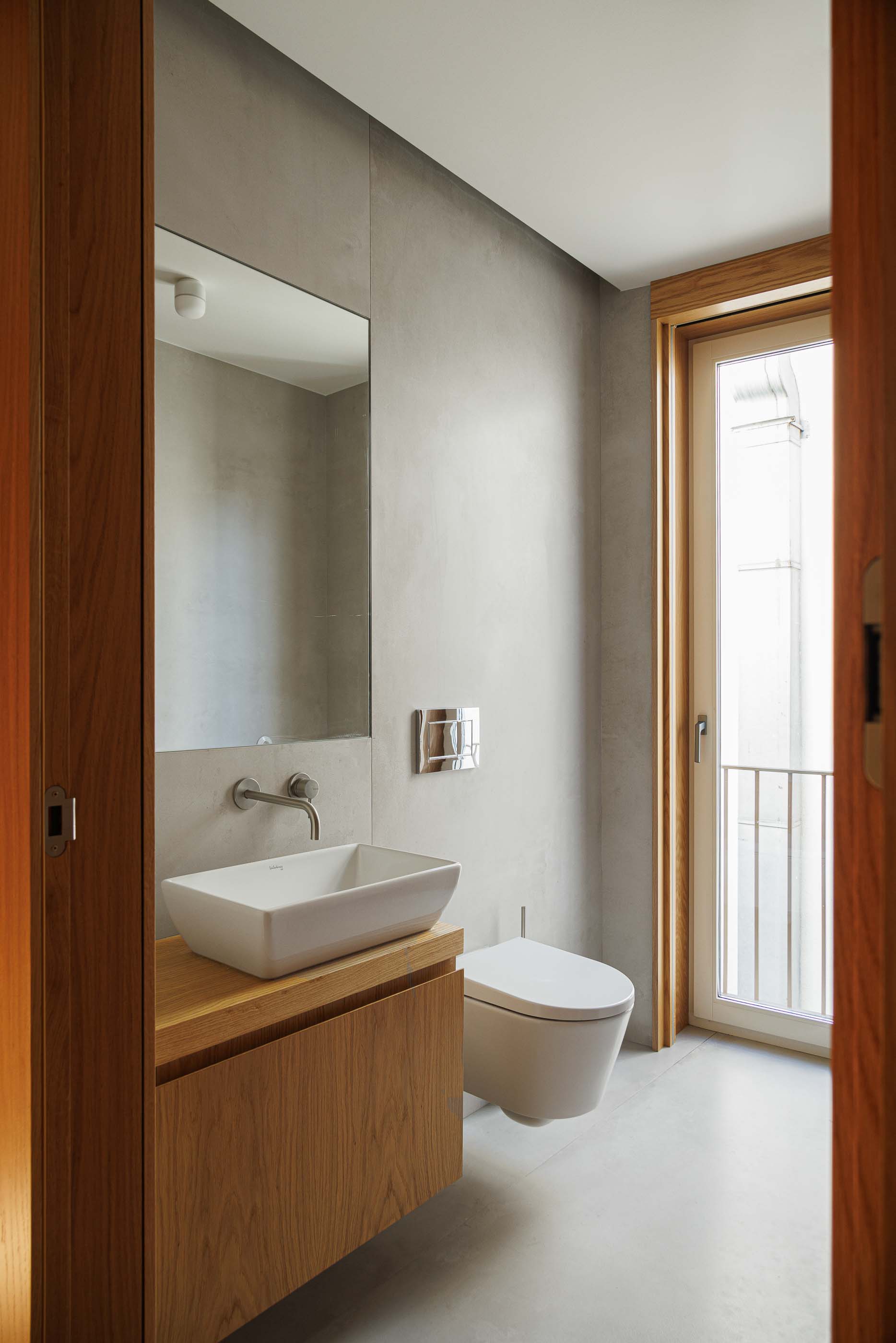 Casa de banho: Lavatório branco sobre móvel em madeira clara, espelho e revestimento cinzento. — Apartamentos Vitória arquitetura Floret Arquitectura em Viseu