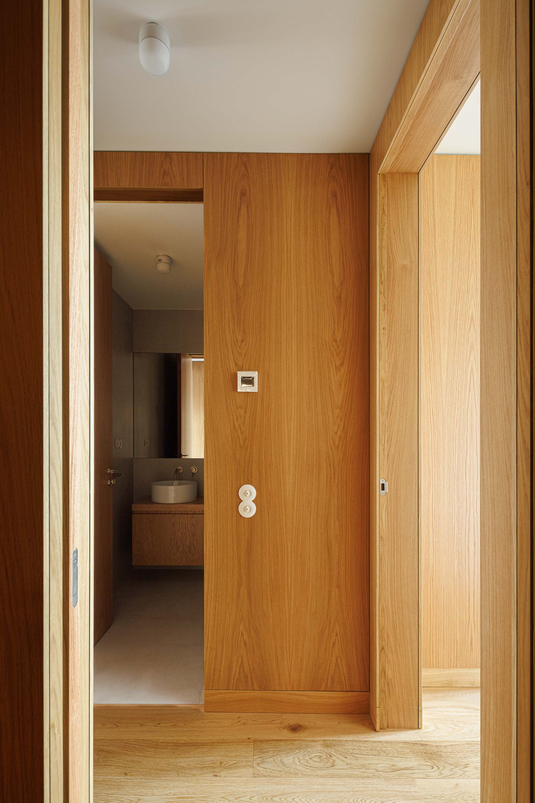 Corredor: Vista para casa de banho com lavatório branco e porta em madeira clara. — Apartamentos Vitória arquitetura Floret Arquitectura em Viseu