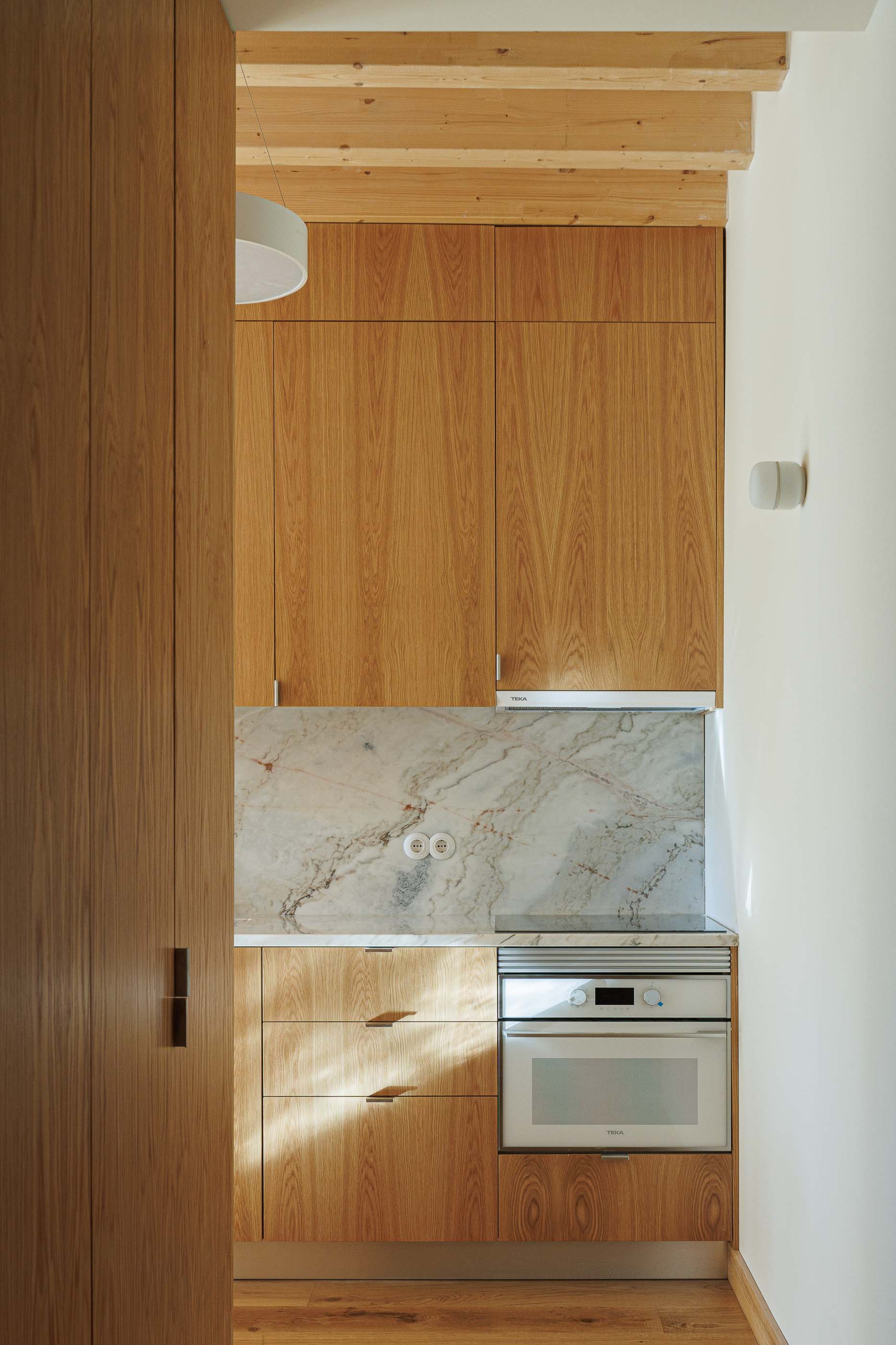 Cozinha: Armários em madeira clara, bancada em mármore e teto com vigas de madeira expostas. — Apartamentos Vitória arquitetura Floret Arquitectura em Viseu