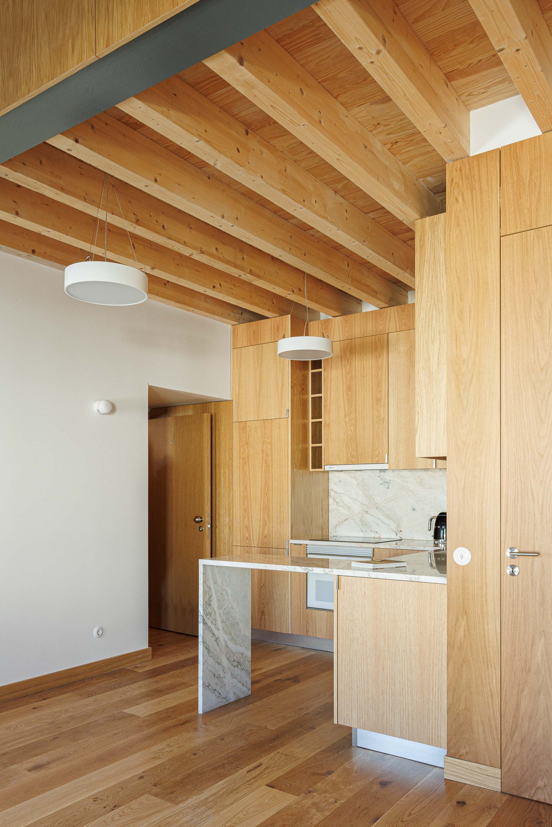 Cozinha: Armários em madeira clara, bancada em mármore e teto com vigas de madeira expostas. — Apartamentos Vitória arquitetura Floret Arquitectura em Viseu