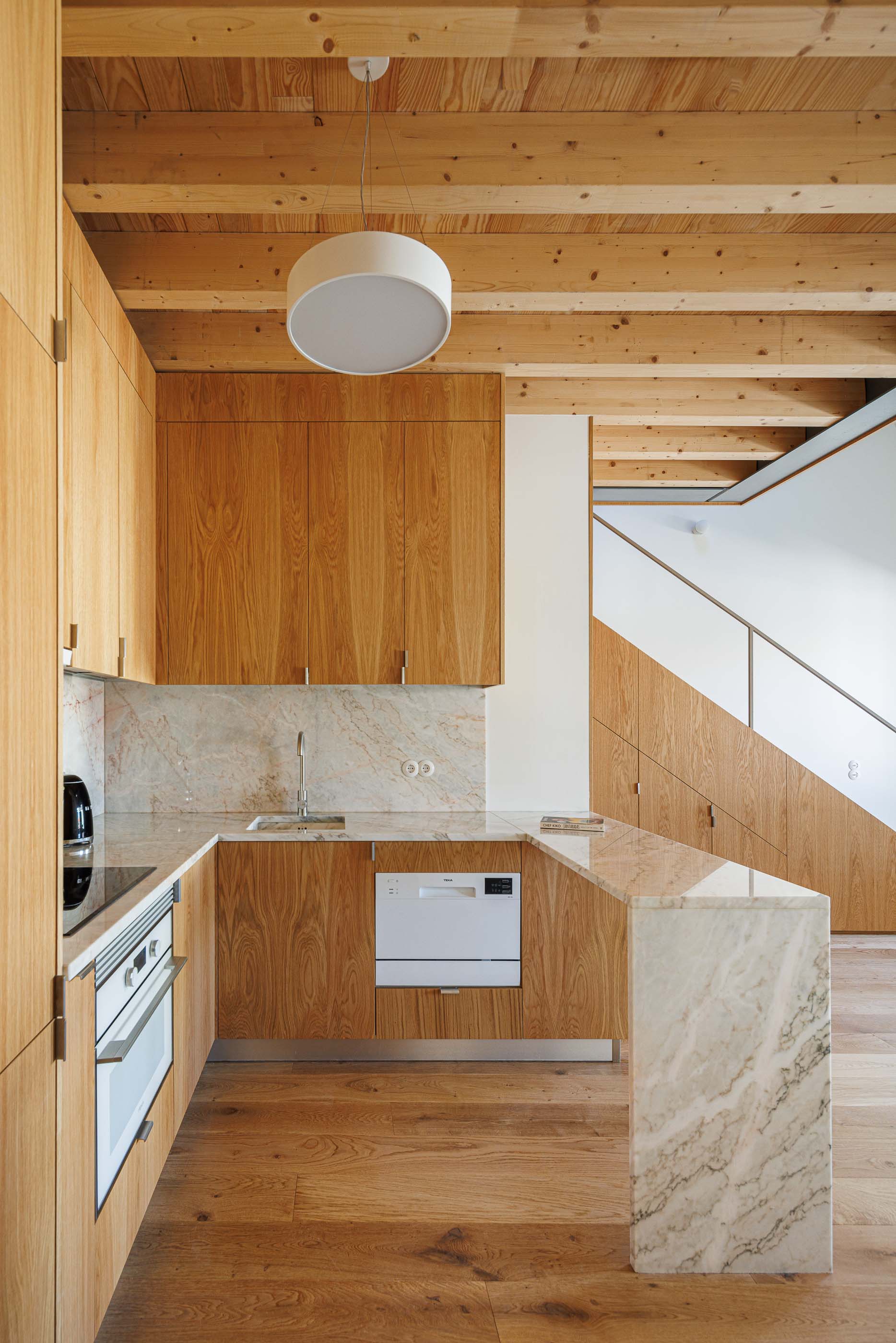 Cozinha: Armários em madeira clara, bancada em mármore e teto com vigas de madeira expostas. — Apartamentos Vitória arquitetura Floret Arquitectura em Viseu