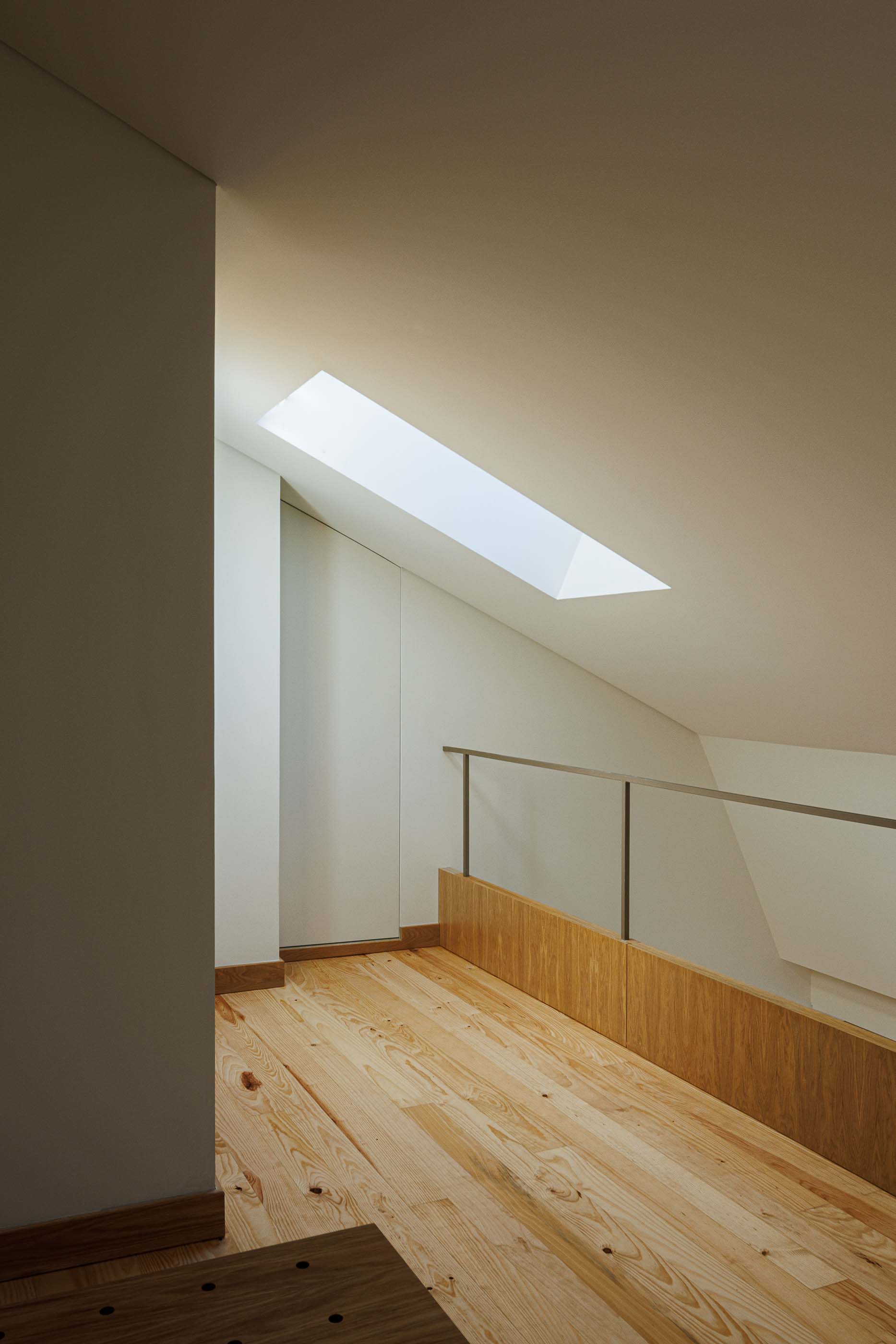 Mezzanine: Sótão com claraboia, pavimento em madeira clara e guarda-corpo metálico. — Apartamentos Vitória arquitetura Floret Arquitectura em Viseu